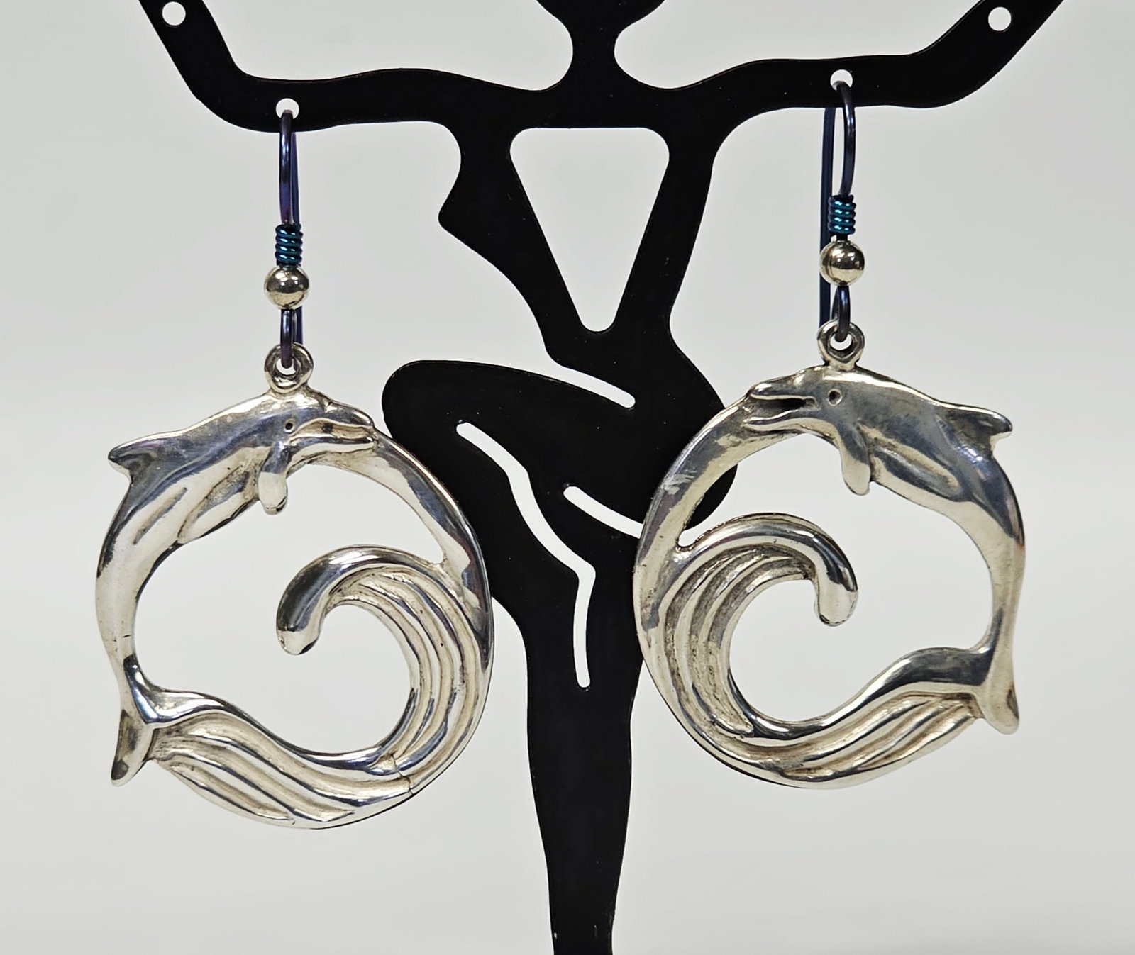 Sterling Silver Dolphin Openwork Earrings 10.8 Grams: A pair of sterling silver drop earrings featuring an openwork circular frame depicting a leaping dolphin over a cresting wave. The earrings are suspended from dark blue-toned shepherd hook ear wires,