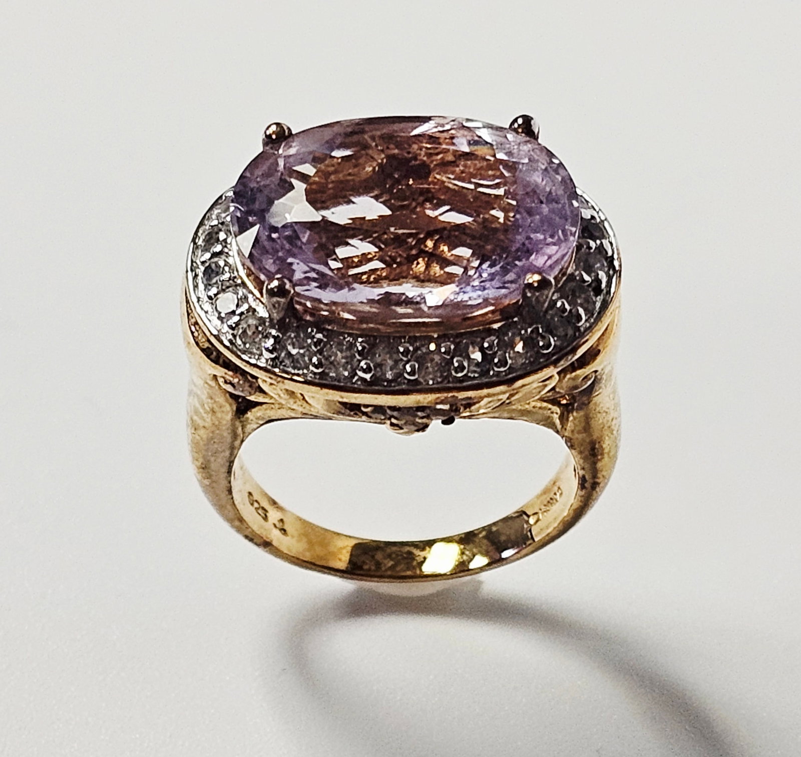 Gold-Plated Sterling Light Purple Gemstone Cocktail Ring 8.4 Grams: A cocktail ring featuring a large, prong-set oval-cut light purple gemstone. The central stone is surrounded by a halo of small, colorless round-cut accent stones. The ornate gallery displays