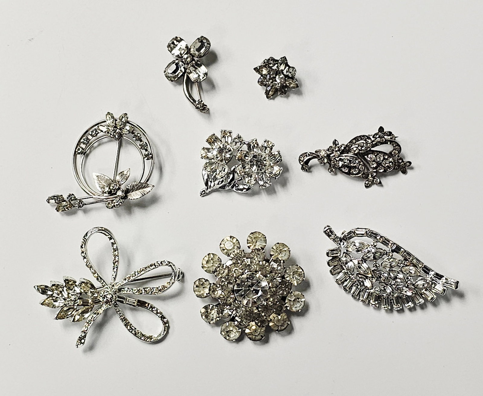 Vintage Rhinestone Costume Jewelry Brooches Inc Krementz: This collection of mid-century costume jewelry features various floral, foliate, and stylized motifs. One circular wreath brooch is composed of a triple-wire frame accented with textured leaves and