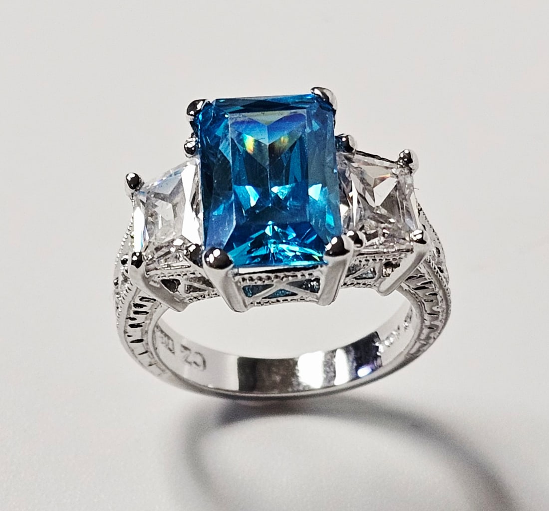 Sterling Silver Blue Cubic Zirconia Cocktail Ring 7.0 Grams: This three-stone cocktail ring is constructed of sterling silver and features a central emerald-cut blue cubic zirconia. The center stone is held in a multi-prong setting and is flanked by two
