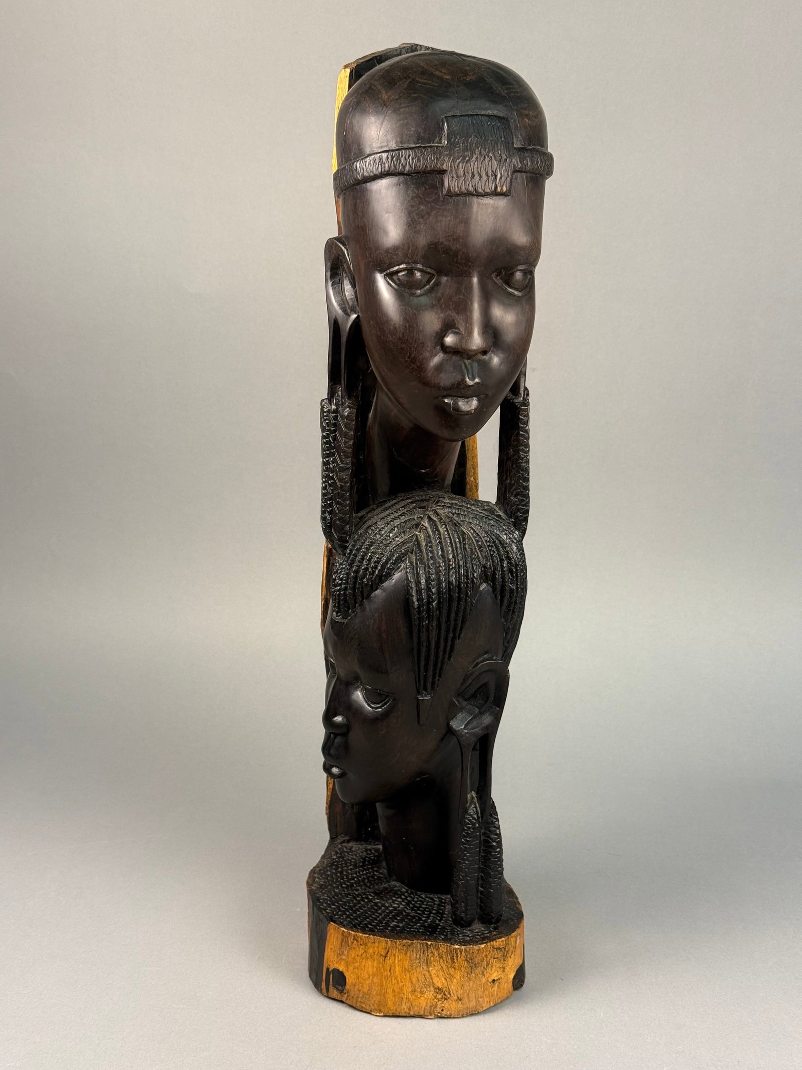 African Hand Carved Ebony Wood Figural Sculpture in the Makonde Style: A vertical figural sculpture hand-carved from a single section of dense hardwood, likely ebony. The piece features two stylized human heads carved in high relief from the dark heartwood, positioned on