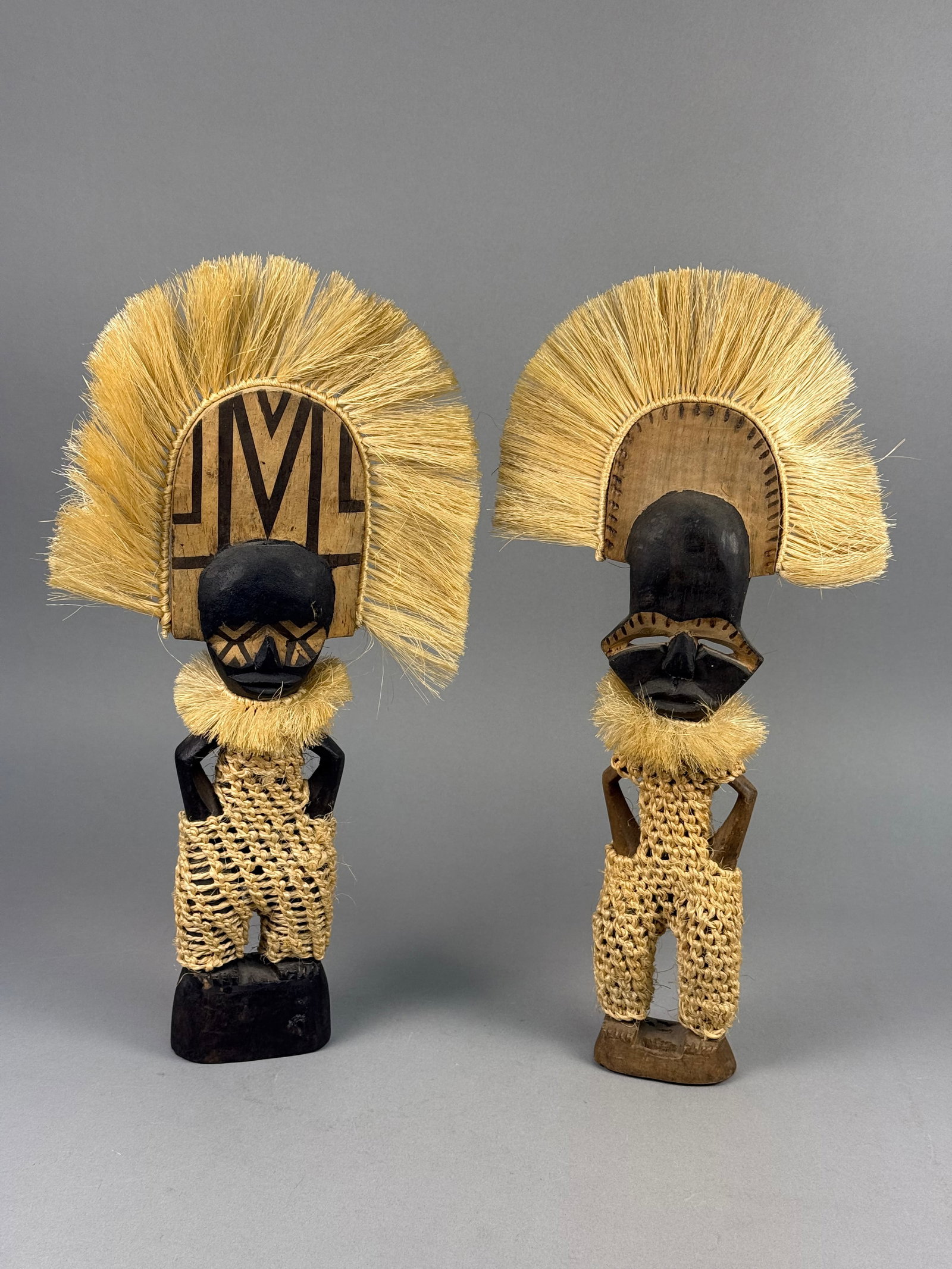 A Pair of Hand-Carved Lozi Style Wooden Sungod Figures with Raffia Headdresses: This pair of hand-carved wooden anthropomorphic figures features natural plant fiber accents and integrated bases. Each figure is carved from a single piece of wood. The heads are topped with semi-cir