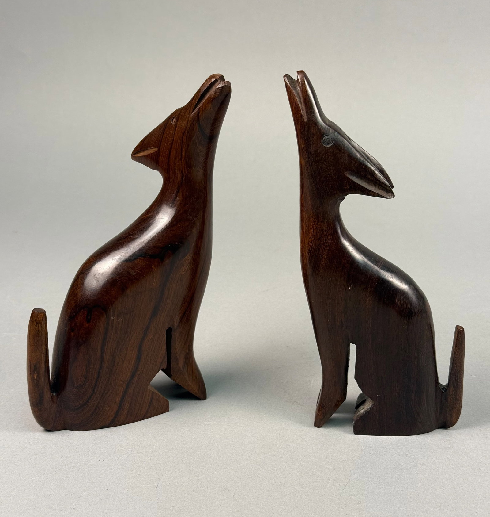 A Pair of Modernist Carved Ironwood Howling Coyote Sculptures: A pair of hand-carved animal sculptures depicting howling coyotes or wolves, crafted from dense, dark ironwood. The figures exhibit a minimalist, modernist aesthetic with elongated necks, stylized ear