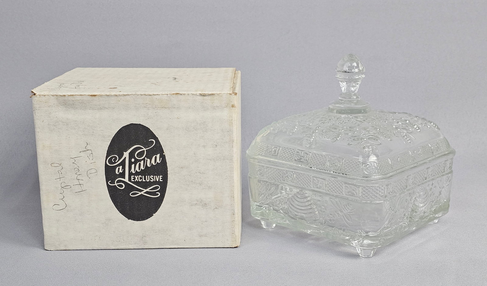 Tiara Exclusives Honey Bee Pattern Clear Pressed Glass Honey Box with Original Box: This clear pressed glass covered dish is distributed by Tiara Exclusives in the Honey Bee pattern, a design frequently produced by the Indiana Glass Company. The square vessel is supported by four mol