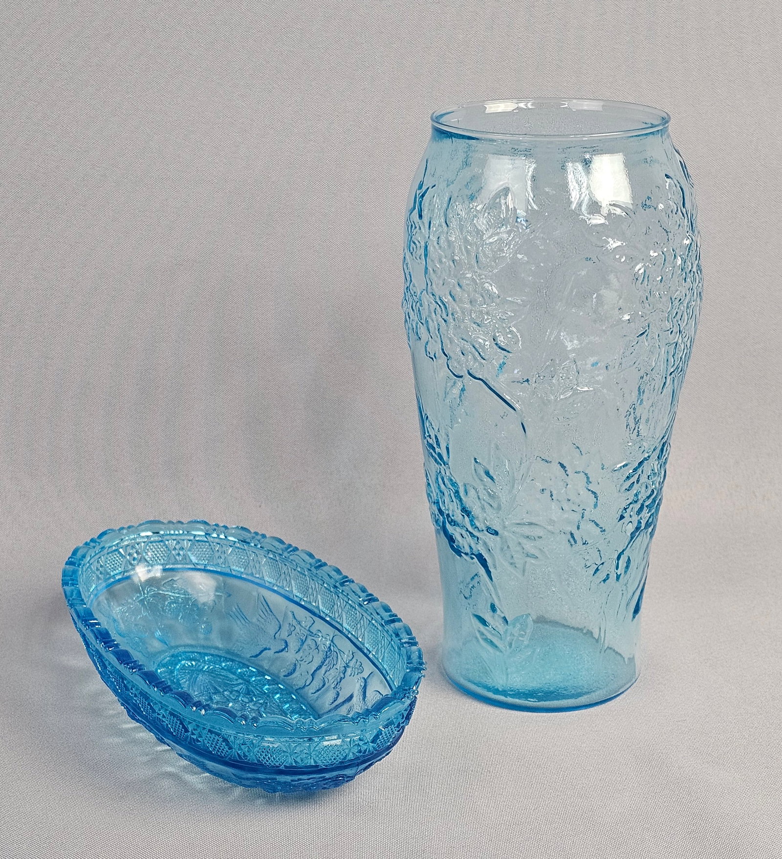 Two Pieces of Blue Pressed Indiana Glass Including a Bird and Strawberry Pattern Bowl: This lot consists of two pieces of blue pressed glass from the early-to-mid 20th century. The first item is a light aqua blue vase featuring a tapering cylindrical form. The exterior is decorated with