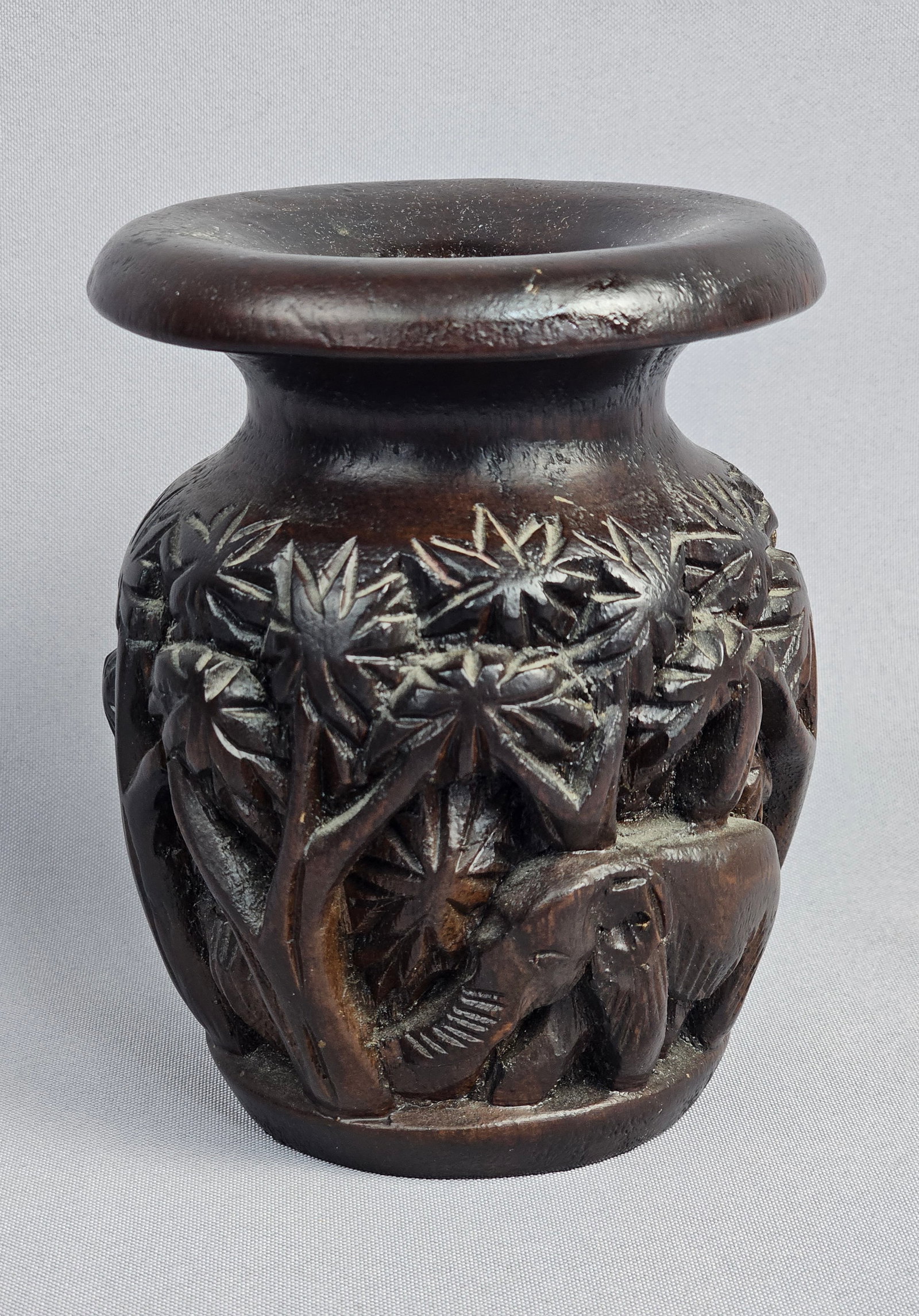 Southeast Asian Hand Carved Hardwood Elephant Motif Vase: This decorative vase is crafted from a dense, dark-stained hardwood. The vessel features a broad, flared, and rounded rim above a short neck, leading to an ovoid body. The exterior is decorated with