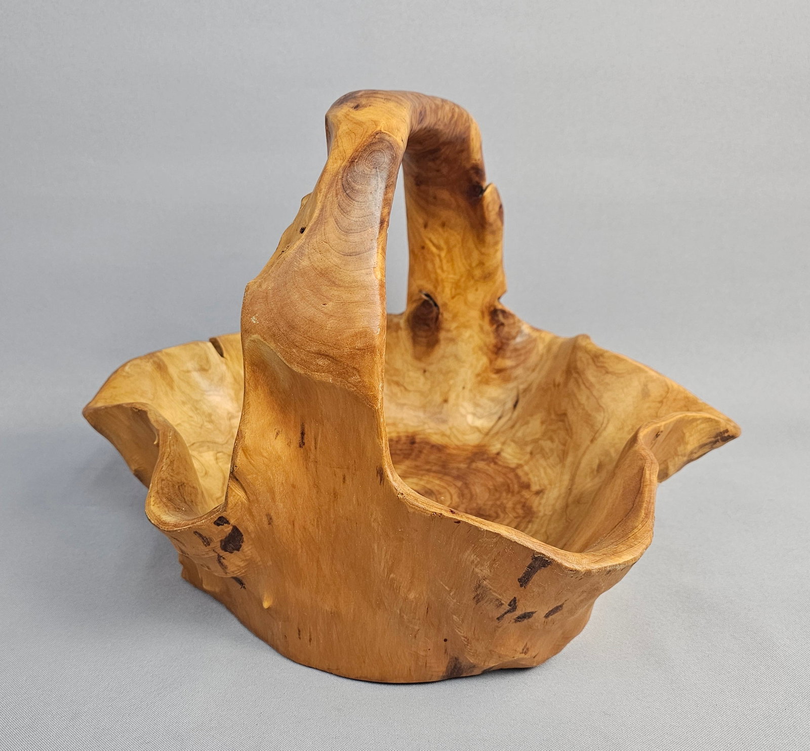 Hand Carved Burl Wood Basket with Integral Handle: A hand-carved rustic basket crafted from a single section of light-toned burl or root wood. The piece features an integral arched handle and an undulating, organic-form rim. The interior and exterior