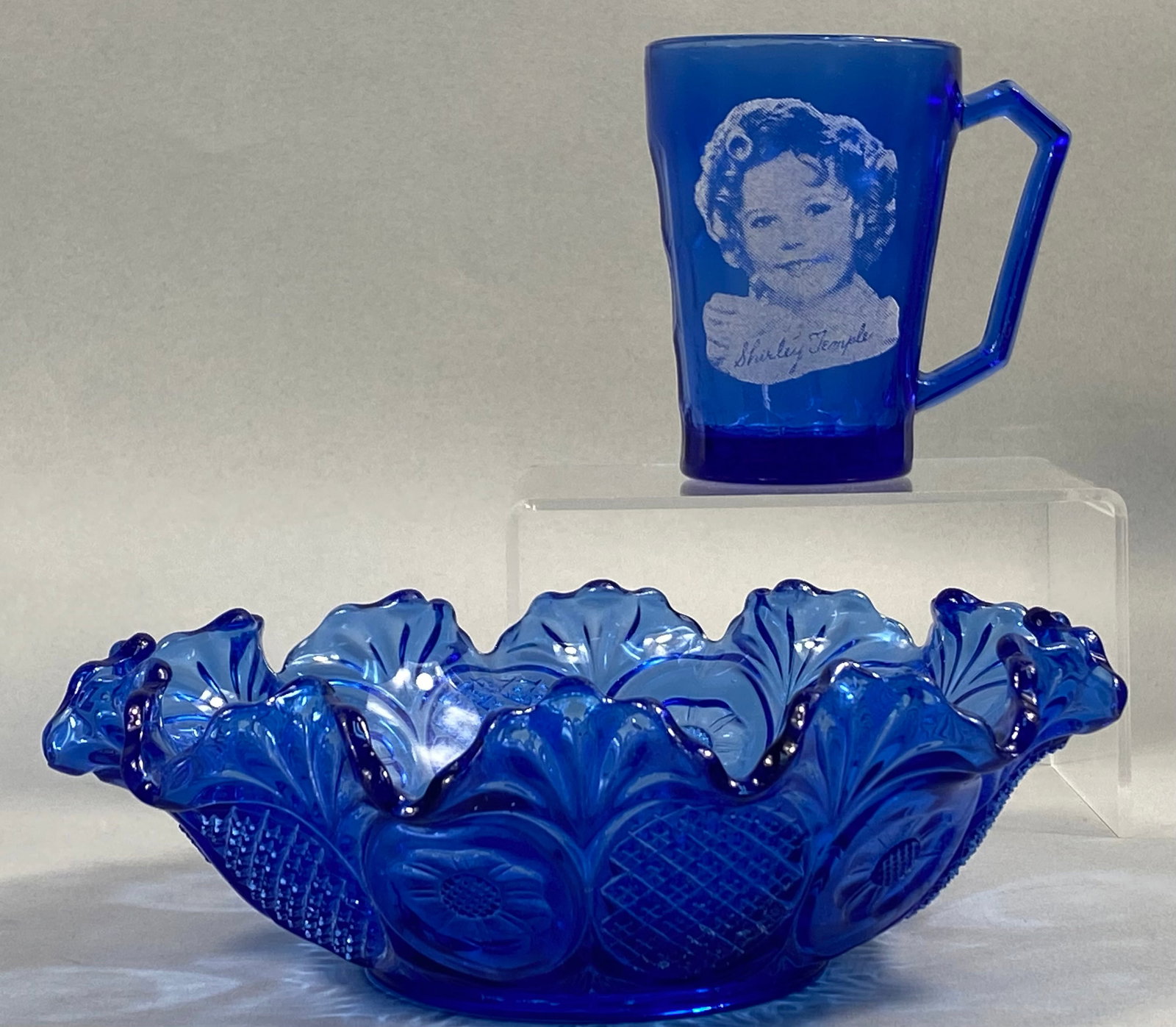 Two Pieces of Cobalt Blue Depression Glass Including Shirley Temple Mug: This lot consists of two pieces of cobalt blue pressed glass characteristic of American production during the Depression era. The first item is a promotional mug featuring a white screen-printed portr