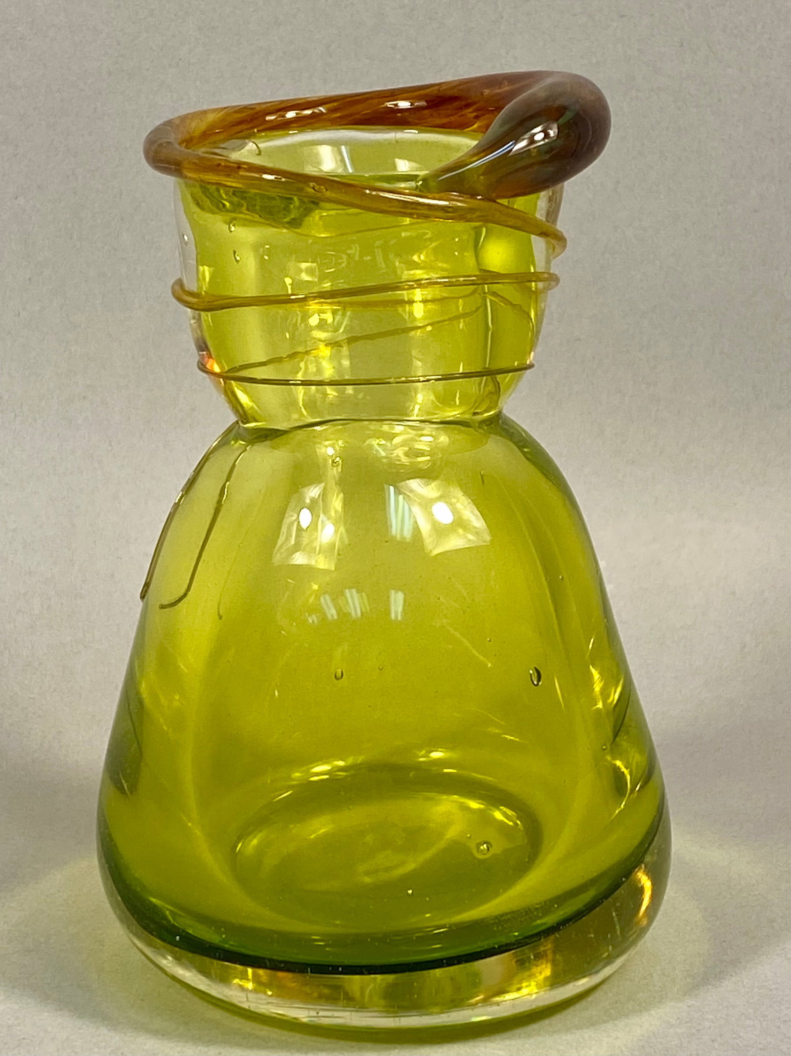 Mid-Century Modern Chartreuse Studio Glass Vase with Applied Amber Trail: This hand-blown studio glass vessel features an organic, waisted form in a translucent chartreuse or lime green hue. The piece is characterized by a thick, applied amber-colored rim with a rounded pro