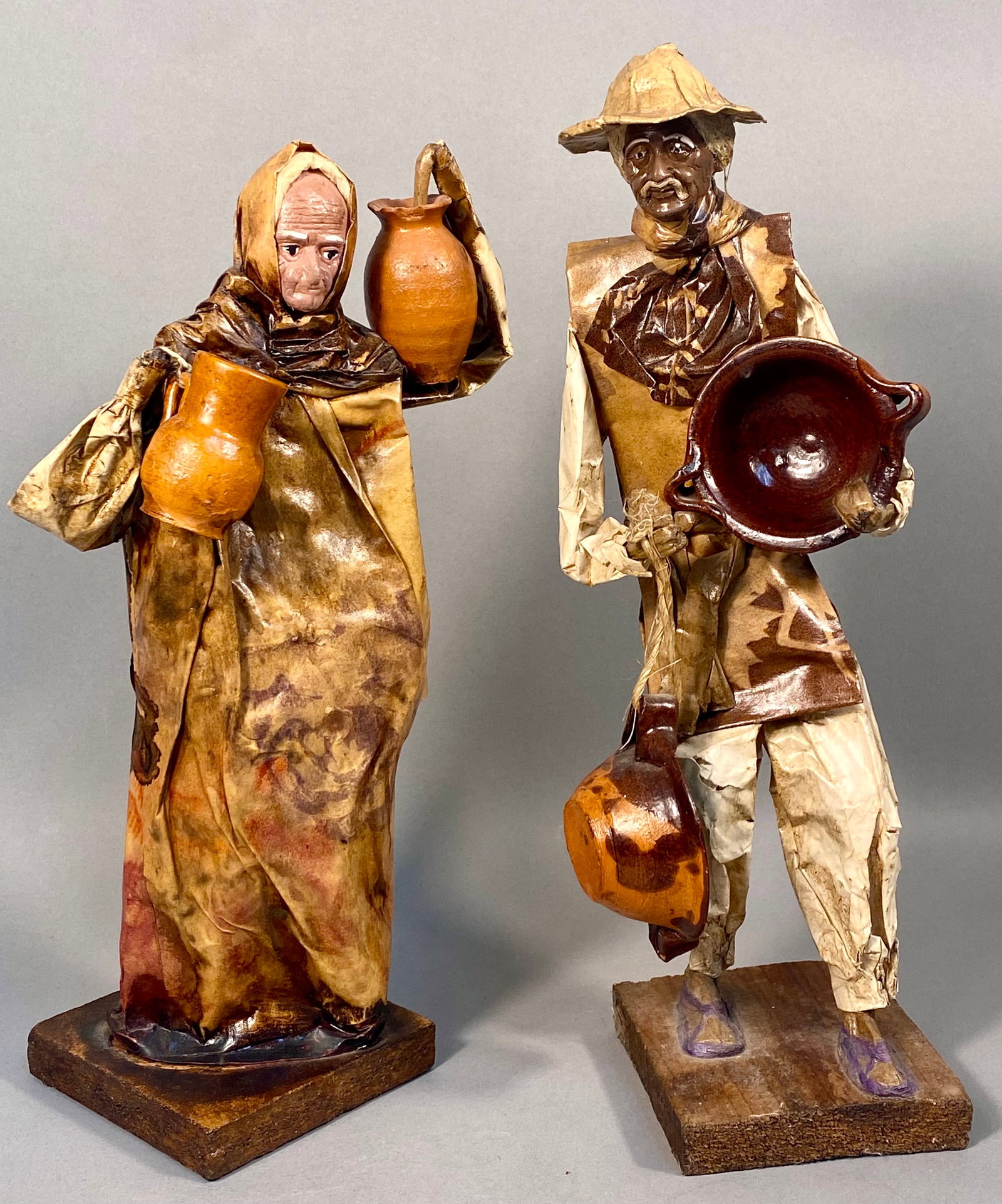A Pair of Mexican Paper Mache Folk Art Figurines with Ceramic Vessels: This pair of handcrafted figurines is attributed as mid-20th-century Mexican folk art. The figures are constructed from stiffened and painted paper or leather over wire armatures, a technique often us