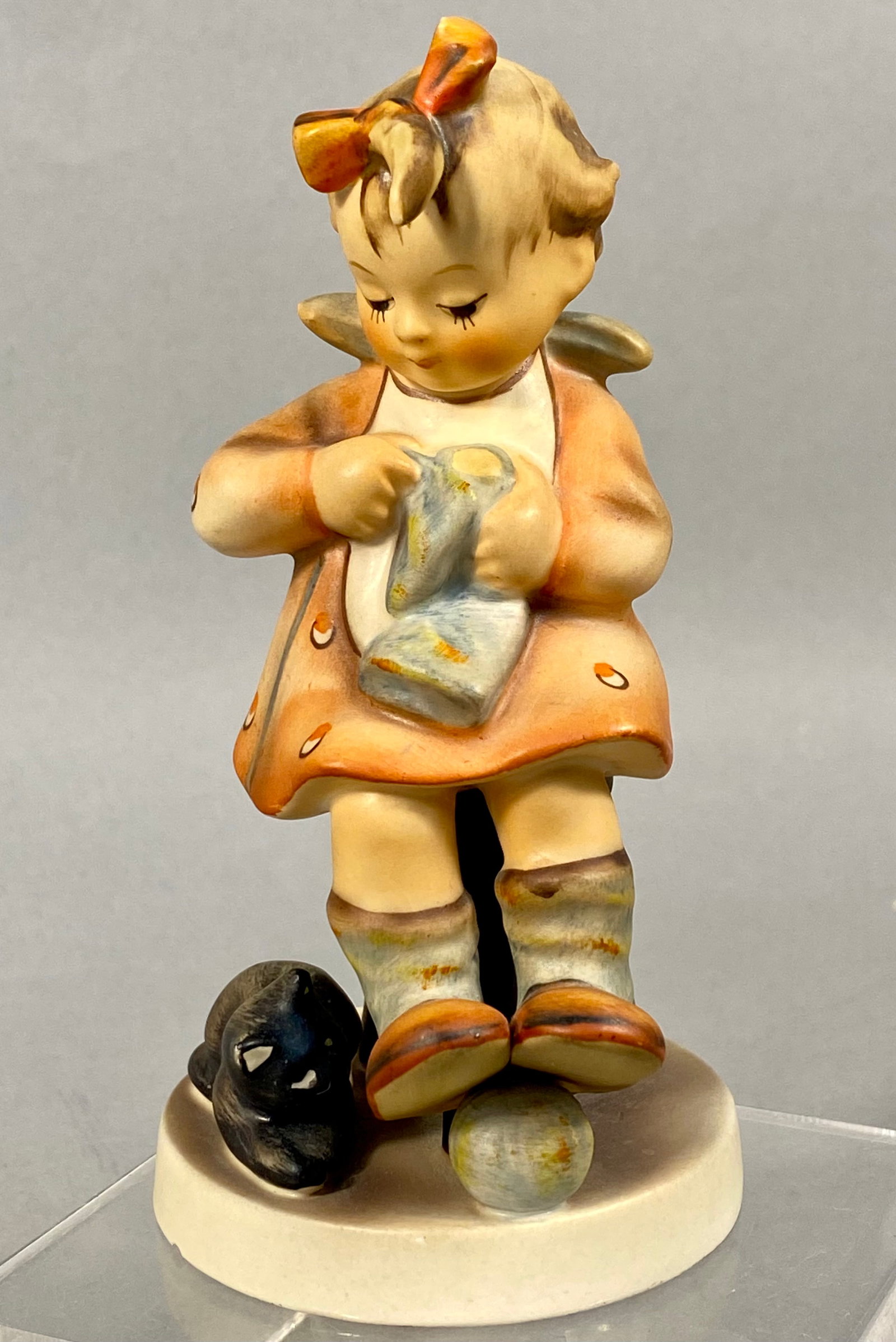 Goebel Hummel Mothers Helper Figurine Number 133 TMK-2: This hand-painted ceramic figurine is a Goebel Hummel titled Mothers Helper, model number 133. The piece depicts a young girl with a red bow in her hair, wearing an orange patterned dress, seated on a