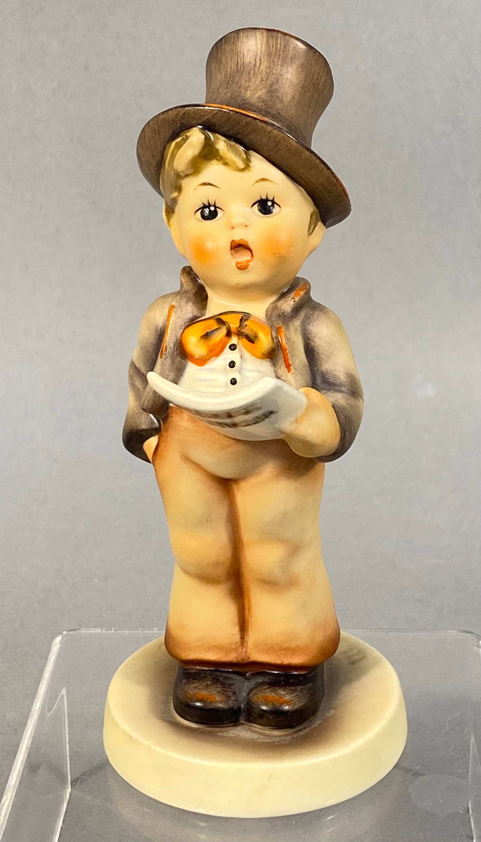 Goebel Hummel Street Singer Figurine Model 131: This Goebel Hummel ceramic figurine is titled Street Singer, model number 131. The hand-painted glazed earthenware figure depicts a young boy wearing a brown top hat, a greyish-tan jacket, a white wai
