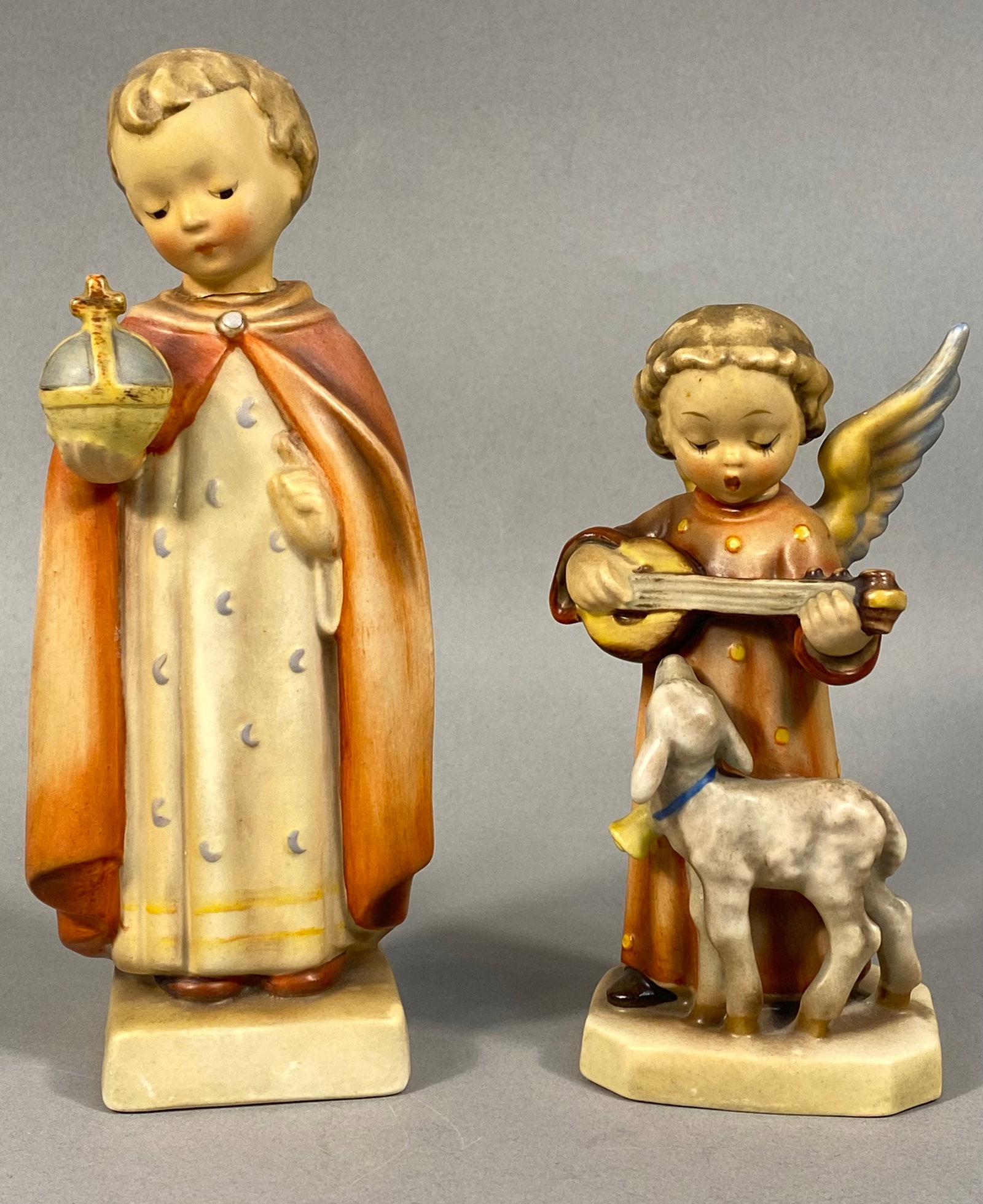 A Pair of Goebel Hummel Figurines, Holy Child and Angel Serenade: A pair of hand-painted ceramic figurines produced by Goebel, featuring designs by Sister Maria Innocentia Hummel. The first figurine, titled Holy Child, depicts the Infant Jesus of Prague wearing a cr