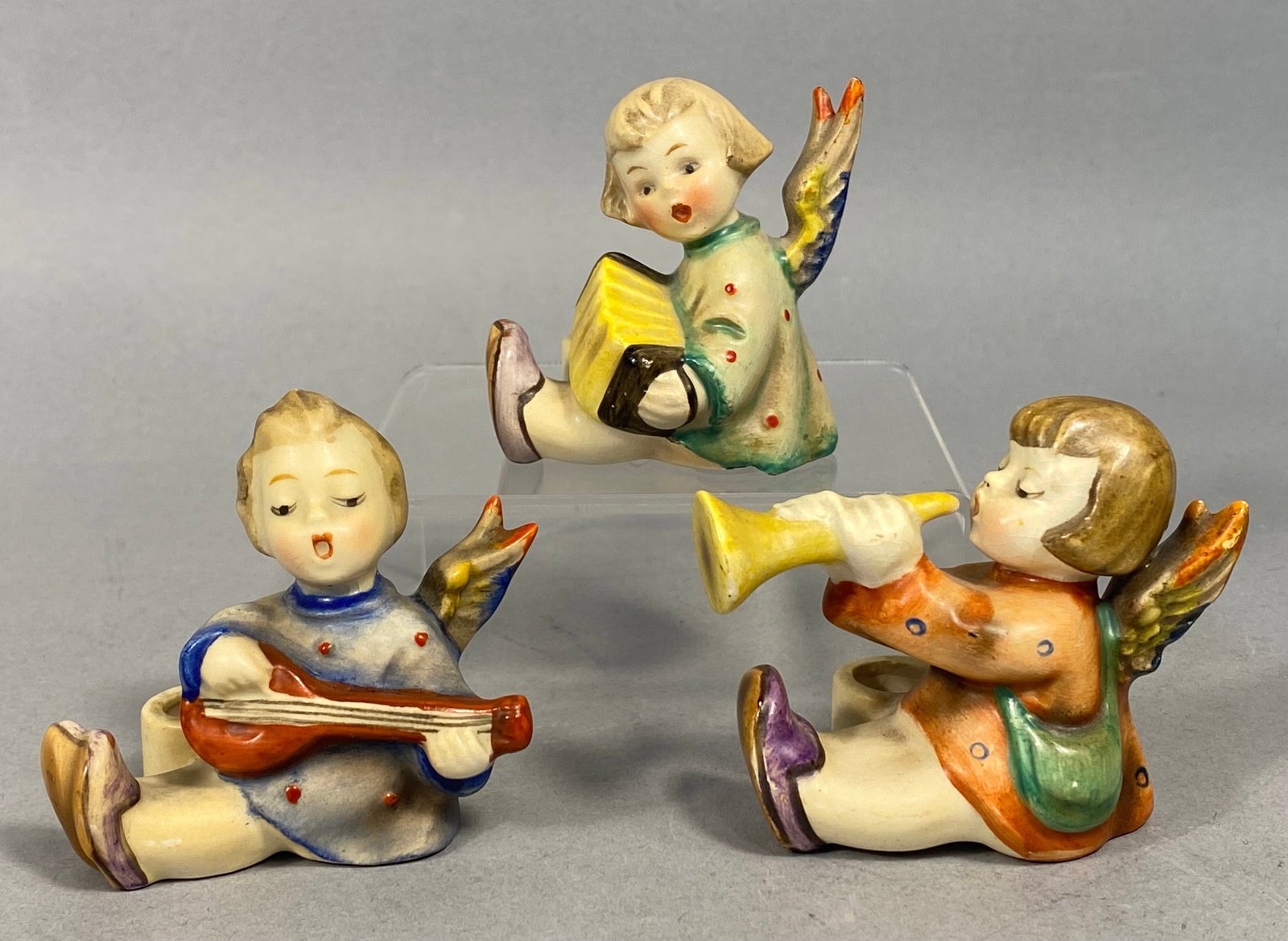 A Trio of Goebel M.I. Hummel Joyous News Angel Candle Holders: This trio of glazed ceramic figurines features seated musical angels from the Joyous News collection, manufactured by W. Goebel Porzellanfabrik and based on the artwork of M.I. Hummel. The set consist