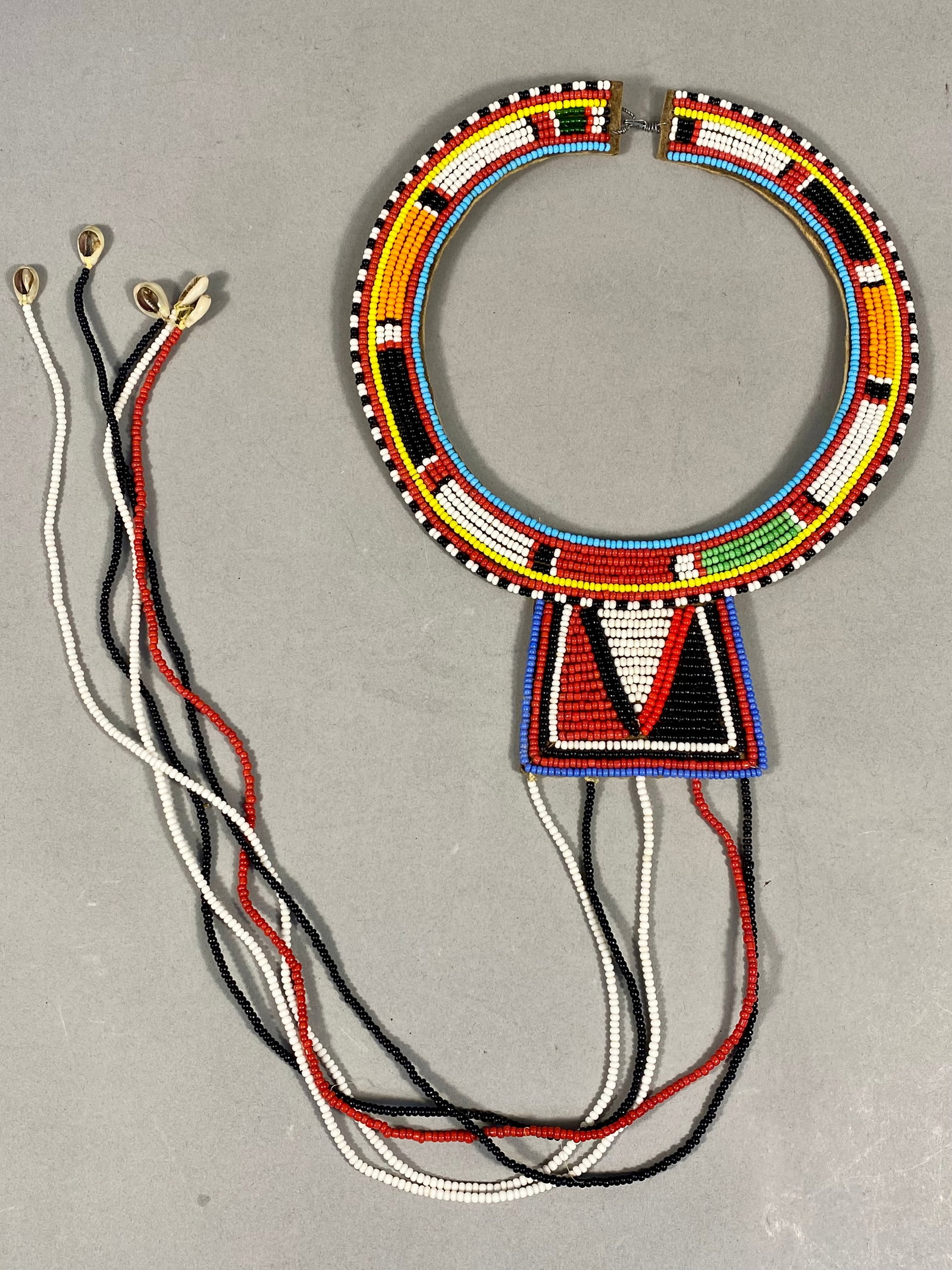 Maasai Beaded Wedding Necklace with Cowrie Shell Tassels: This Maasai beaded wedding necklace features a circular collar and a central rectangular bib-style pendant. The piece is constructed with multicolored glass seed beads in red, white, black, yellow, or