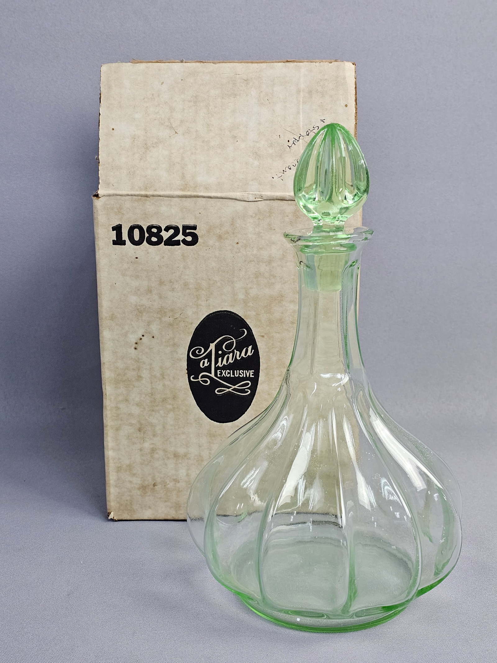 Tiara Exclusives Light Green Ribbed Glass Decanter with Stopper and Original Box: This vintage pressed glass decanter is a product of Tiara Exclusives. The vessel is crafted from light green translucent glass and features a flared, multi-paneled ribbed body that tapers to a slender