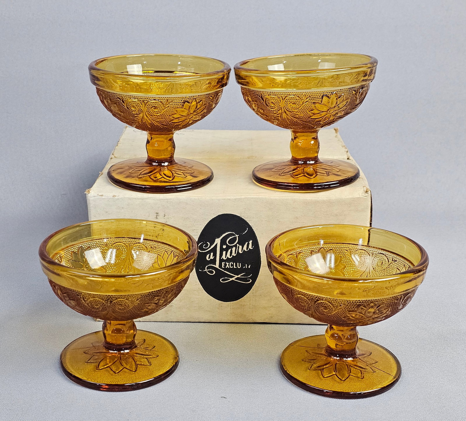 Set of Four Indiana Glass Tiara Sandwich Pattern Amber Sherbet Dishes: This set of four stemmed sherbet dishes is constructed from amber-colored pressed glass. The pieces feature the Sandwich pattern, which is defined by an intricate floral and scroll motif set against a