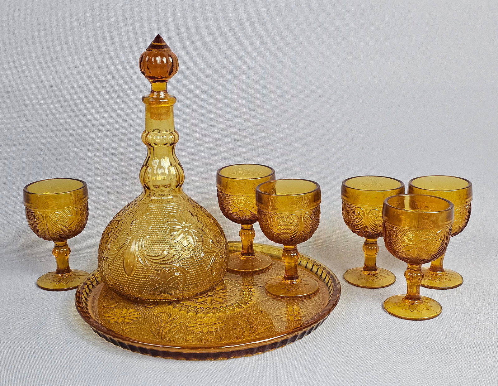 Tiara Exclusives Amber Sandwich Pattern Pressed Glass Decanter Set with Original Box: This eight-piece pressed glass set is produced in the Sandwich pattern, featuring relief-molded daisy and scrolling foliage motifs against a stippled background. The set is executed in a translucent a
