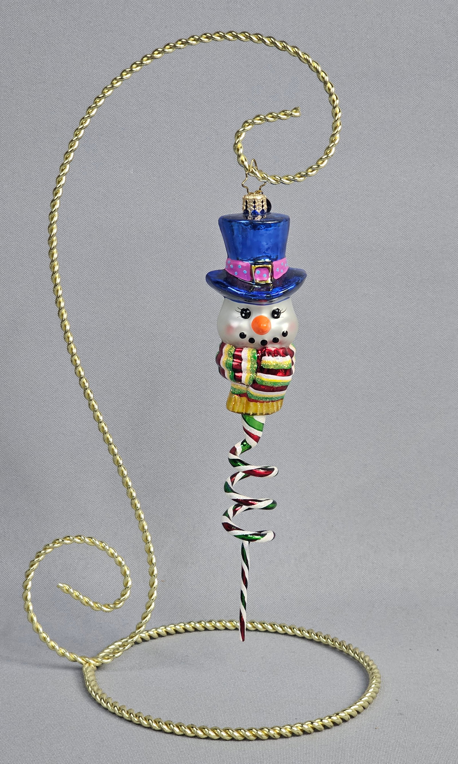 Christopher Radko Hand-Blown Glass Snowman Ornament with Spiral Tail and Stand: Stand not included. A hand-blown glass Christmas ornament produced by Christopher Radko. The ornament features a stylized snowman figure wearing a translucent cobalt blue top hat accented with a pink 