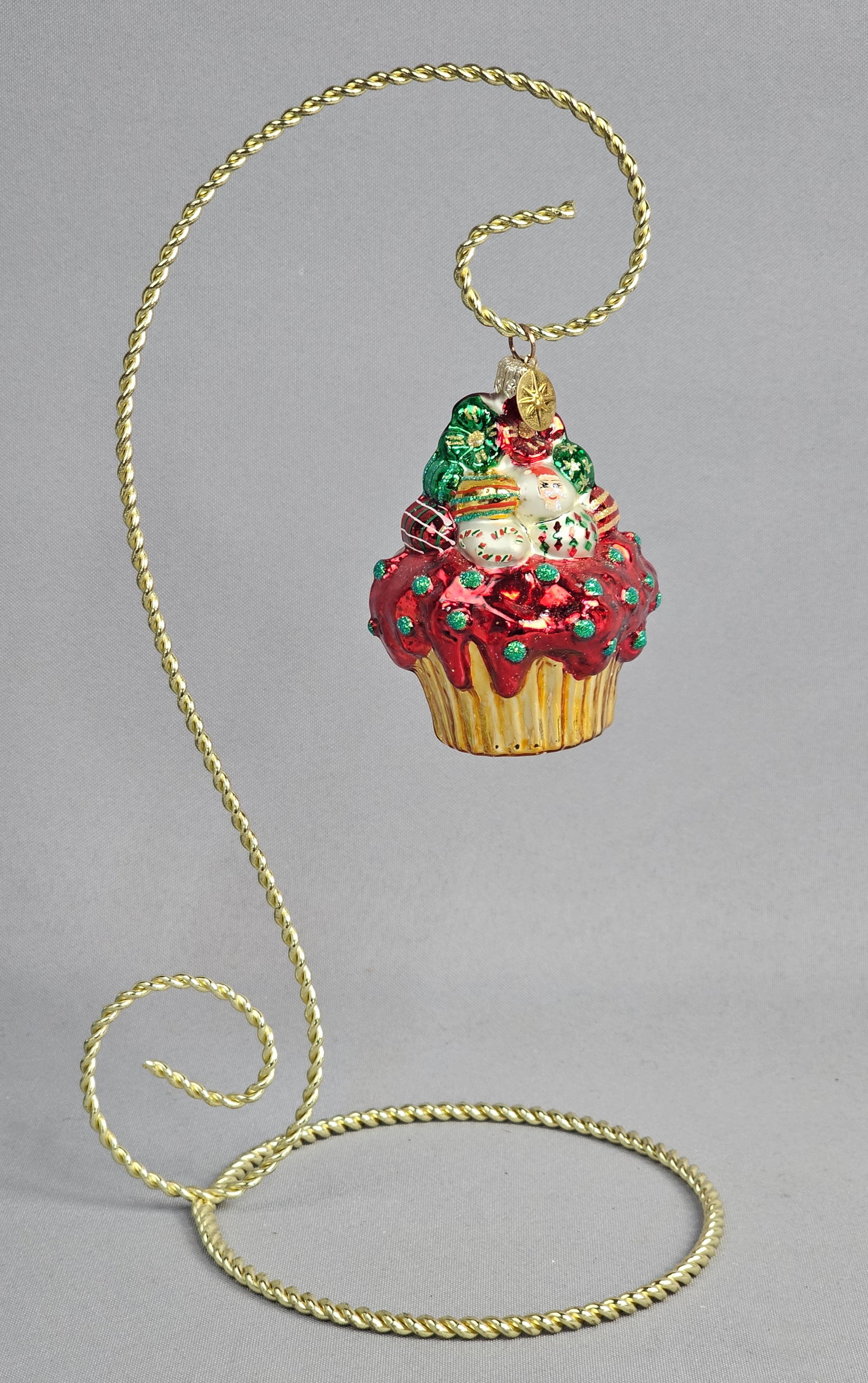 Old World Christmas Blown Glass Holiday Cupcake Ornament: This mouth-blown glass Christmas ornament is produced by Old World Christmas and is designed in the form of a holiday-themed cupcake. The ornament features a gold-toned metallic fluted base representi