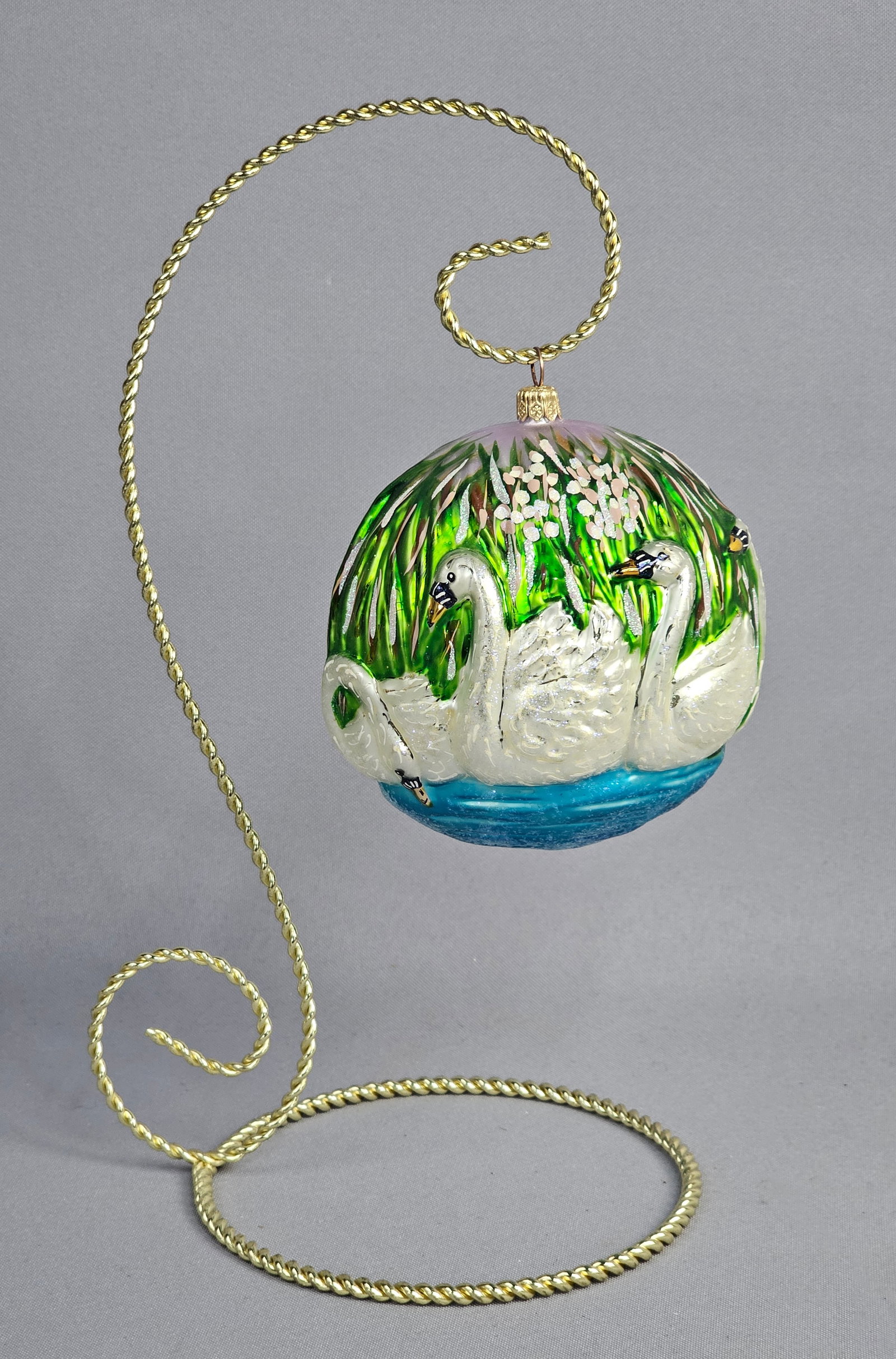 Komozja Polish Hand-Painted Glass Swan Scene Christmas Ornament with Display Stand: A mouth-blown glass Christmas ornament manufactured by Komozja in Poland. The spherical orb features a scenic, hand-painted depiction of three white swans swimming in a pond rendered in blue. The land