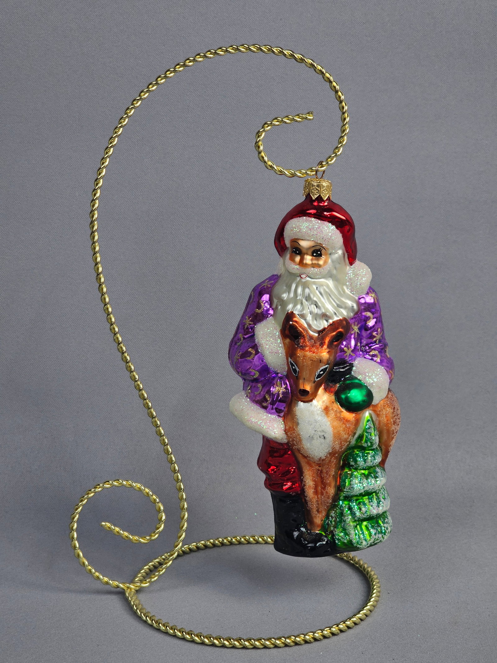 Christopher Radko Glass Santa with Reindeer and Evergreen Tree Christmas Ornament: A mold-blown glass figural Christmas ornament by Christopher Radko depicting Santa Claus holding a brown reindeer fawn and a small green evergreen tree. Santa is portrayed wearing a non-traditional vi