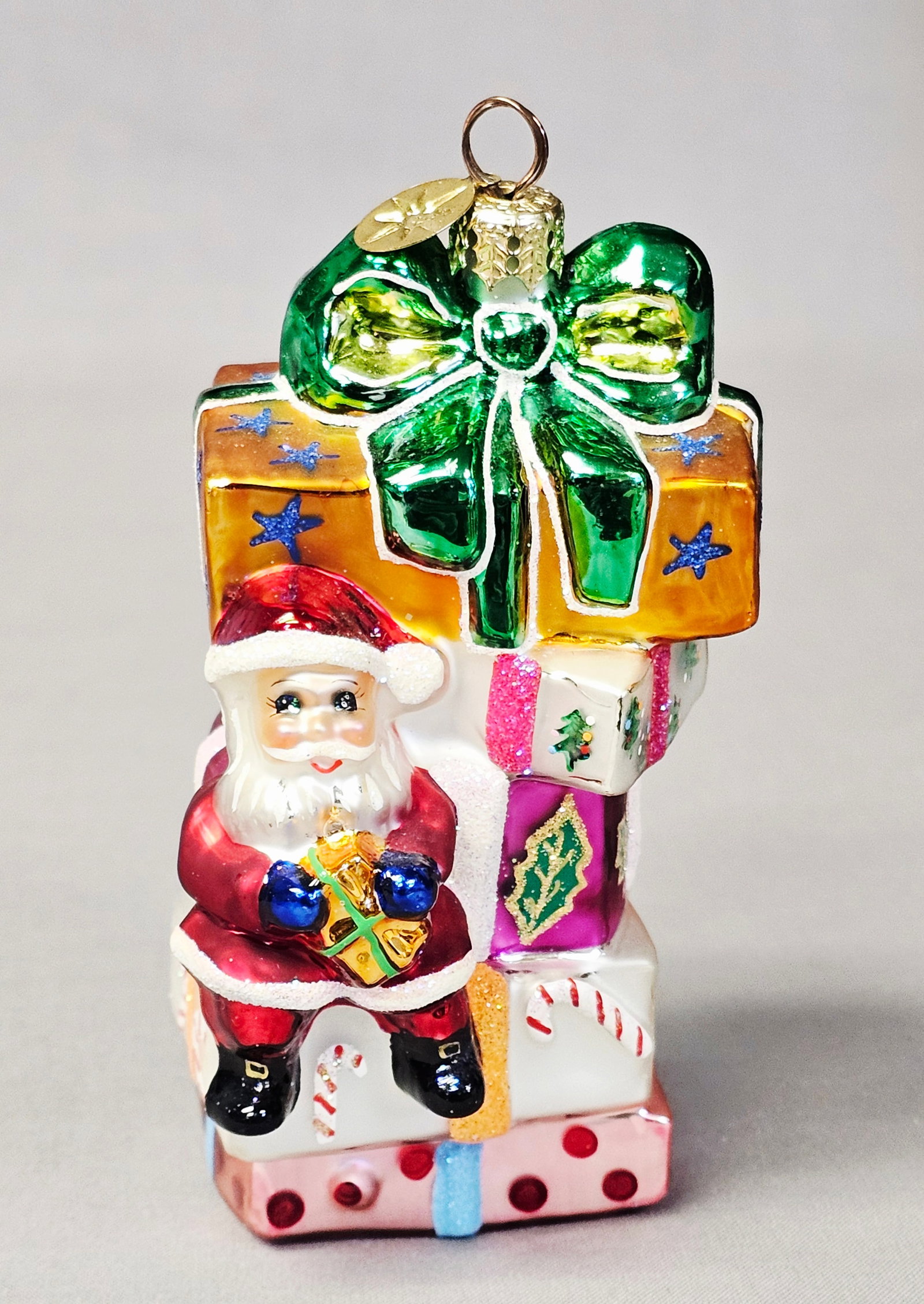 Christopher Radko Glass Santa & Gifts Christmas Ornament: This Christopher Radko mouth-blown glass Christmas ornament features the signature gold-tone eight-pointed starburst charm attached to the hanging loop. The ornament depicts a vertical stack of wrappe