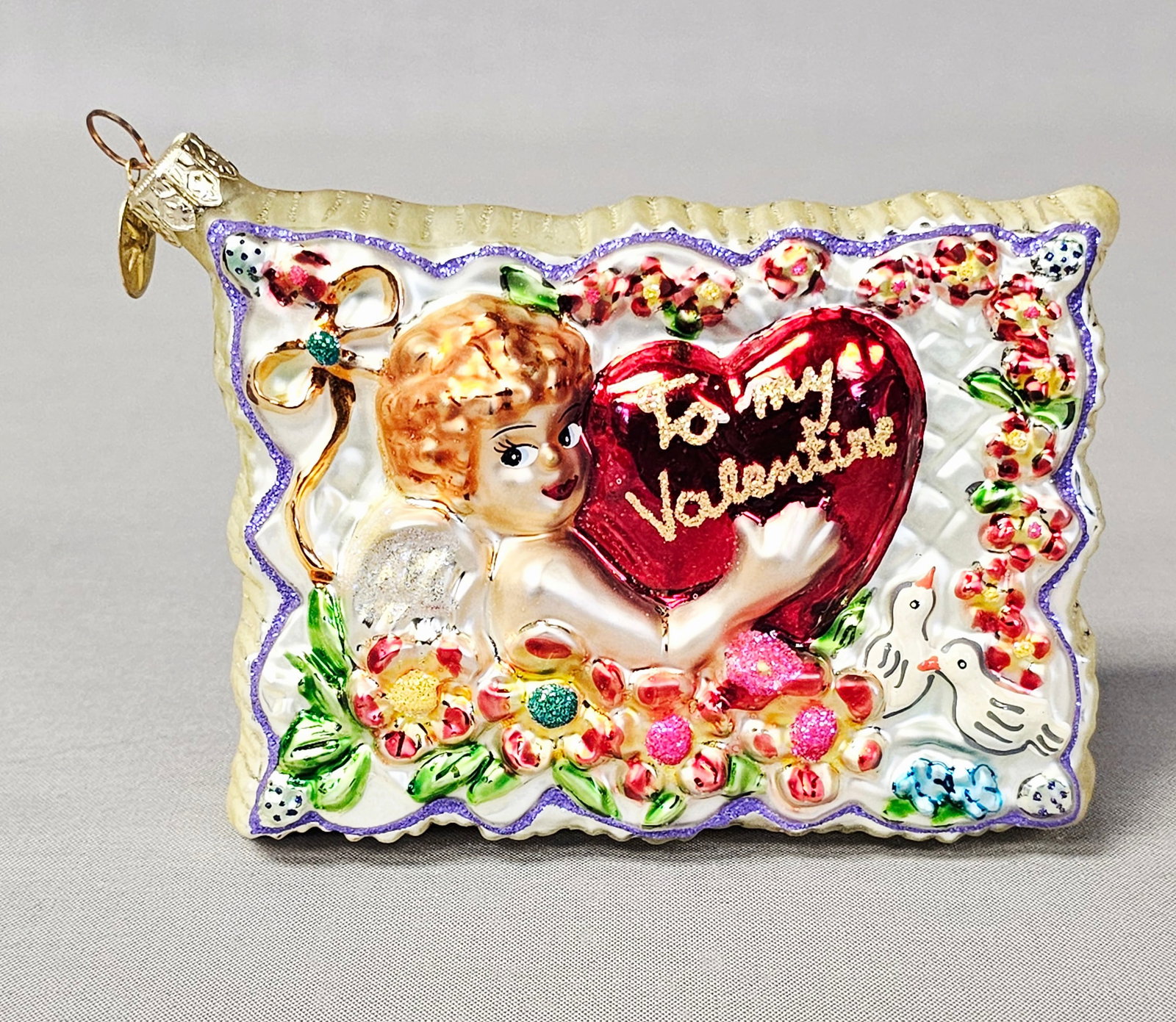 Christopher Radko To My Valentine Glass Postcard Ornament: Stand not included. A Christopher Radko hand-blown glass holiday ornament in a rectangular pillow form with a dual-sided design. The obverse features a relief scene of a winged cherub holding a large 