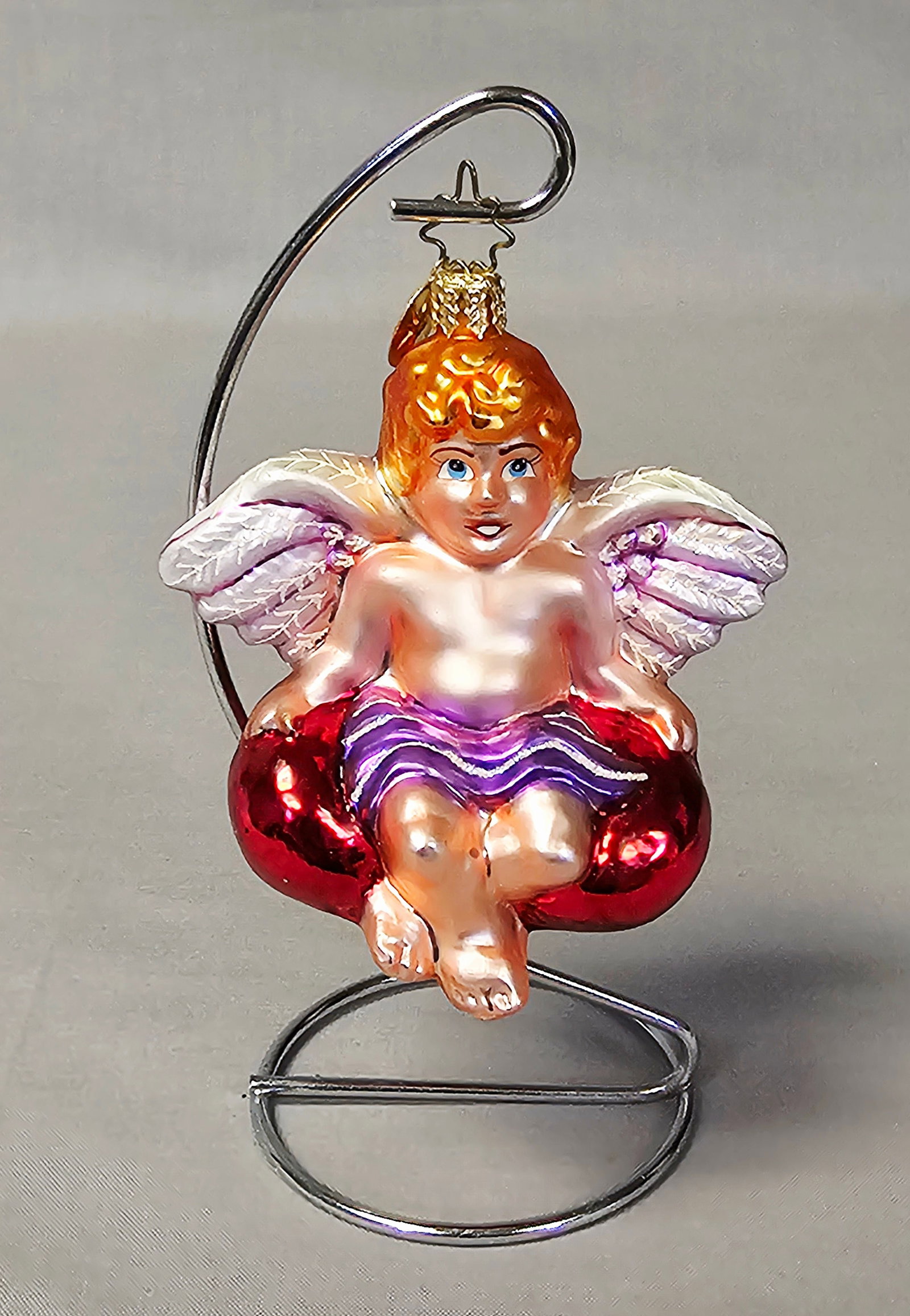 Christopher Radko Glass Cherub on Heart Ornament: Stand not included. This Christopher Radko hand-blown glass ornament depicts a seated cherub or Cupid figure resting upon a large, glossy red heart. The ornament is in excellent condition. There are n