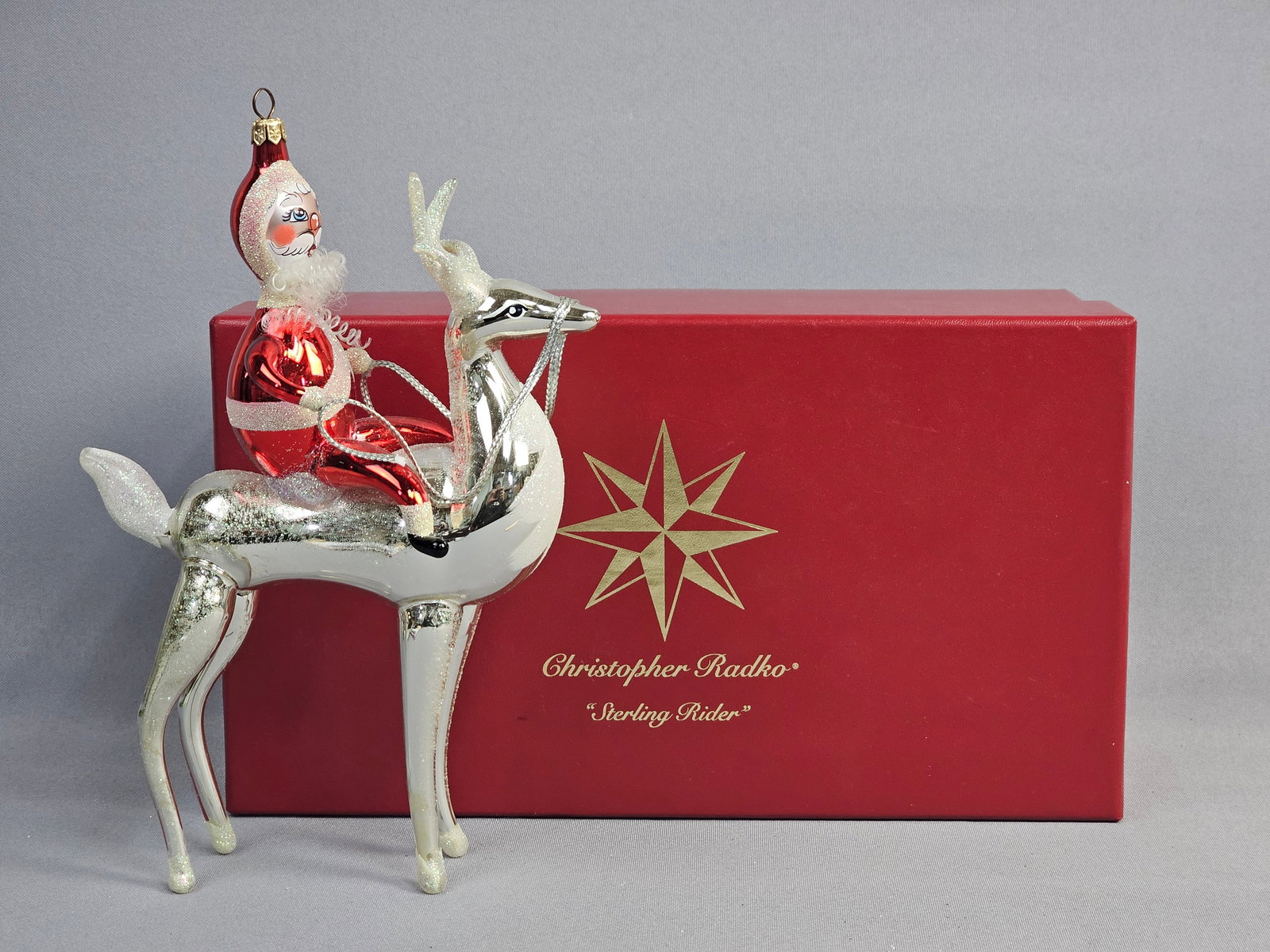 Christopher Radko Sterling Rider Hand-Blown Glass Christmas Ornament with Original Box: This hand-blown glass Christmas ornament, titled Sterling Rider, is produced by designer Christopher Radko. The ornament depicts Santa Claus in a glossy red suit with white glitter trim riding a silve