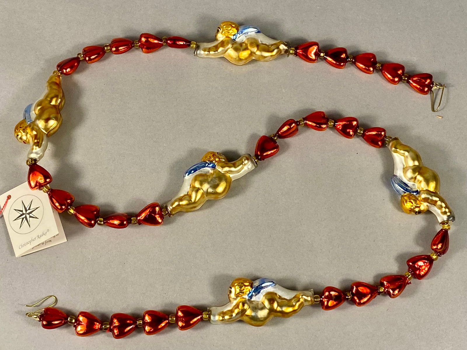 Radko Cupid Flight Hand Blown Glass Garland: A Christopher Radko Cupid Flight holiday garland featuring five hand-blown glass cherub figures and red heart-shaped beads. The cherub figures are decorated with metallic gold, blue, and white lacquer