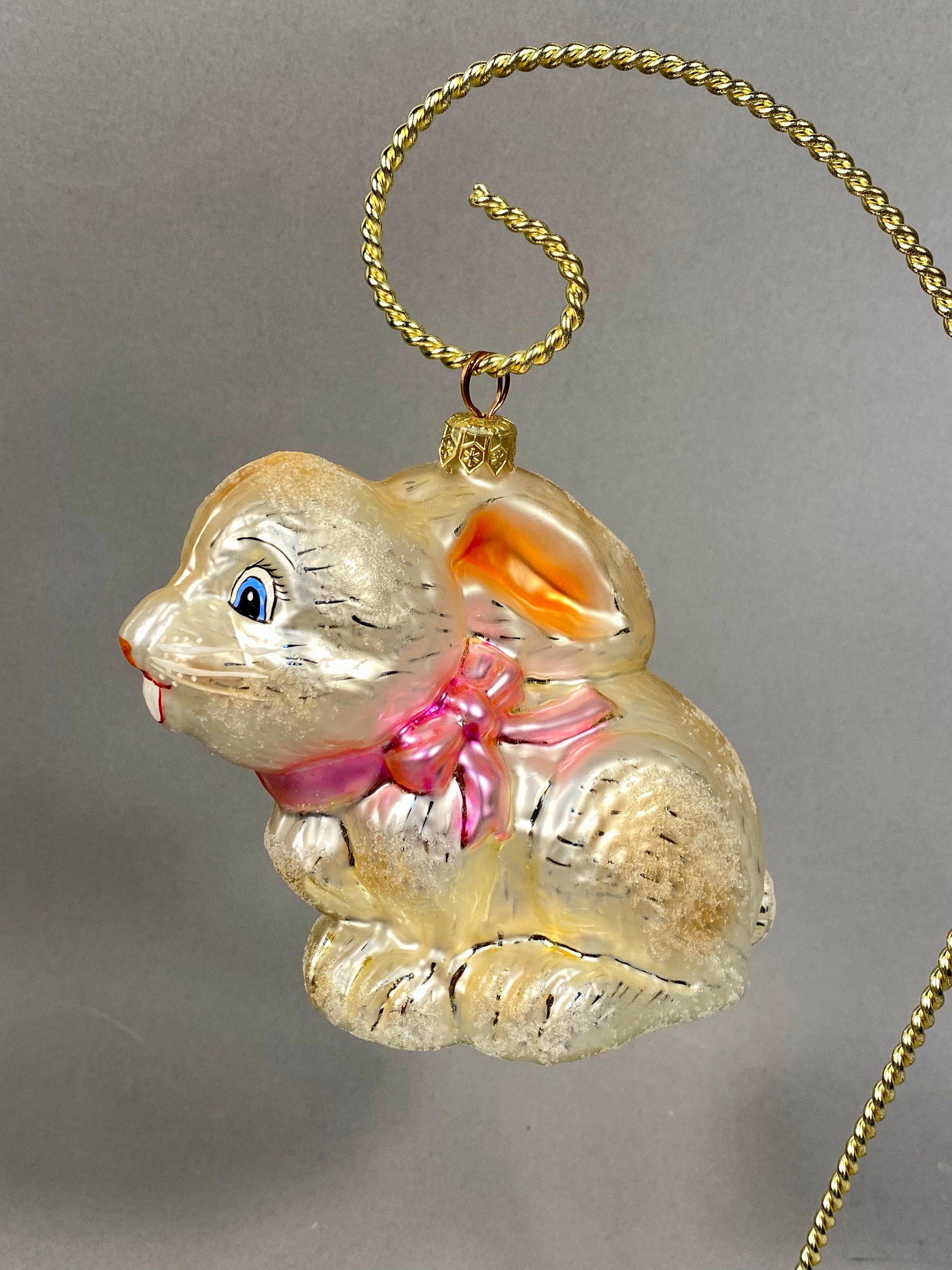 Radko Blown Glass Seated Rabbit Ornament: The ornament features a pearlescent champagne-colored finish with a silvered interior. Hand-painted details include bright blue eyes, a vibrant pink bow tied around the neck, and delicate dark lines s