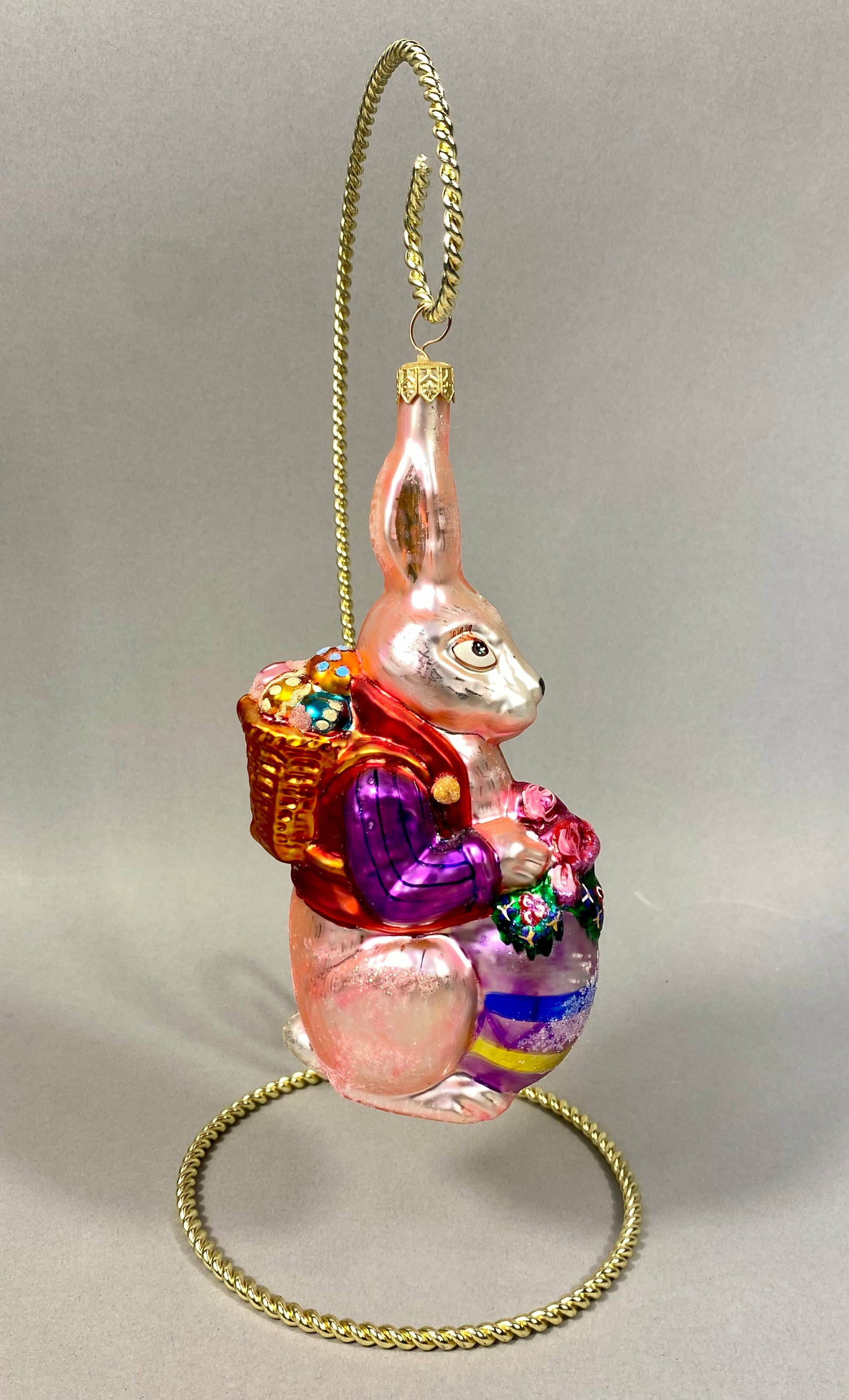 Radko Roy Rabbit Hand-Blown Glass Easter Ornament: The ornament features an anthropomorphic rabbit with an iridescent pink body and hand-painted facial features. The rabbit is dressed in a red vest over a magenta and purple striped long-sleeved shirt.