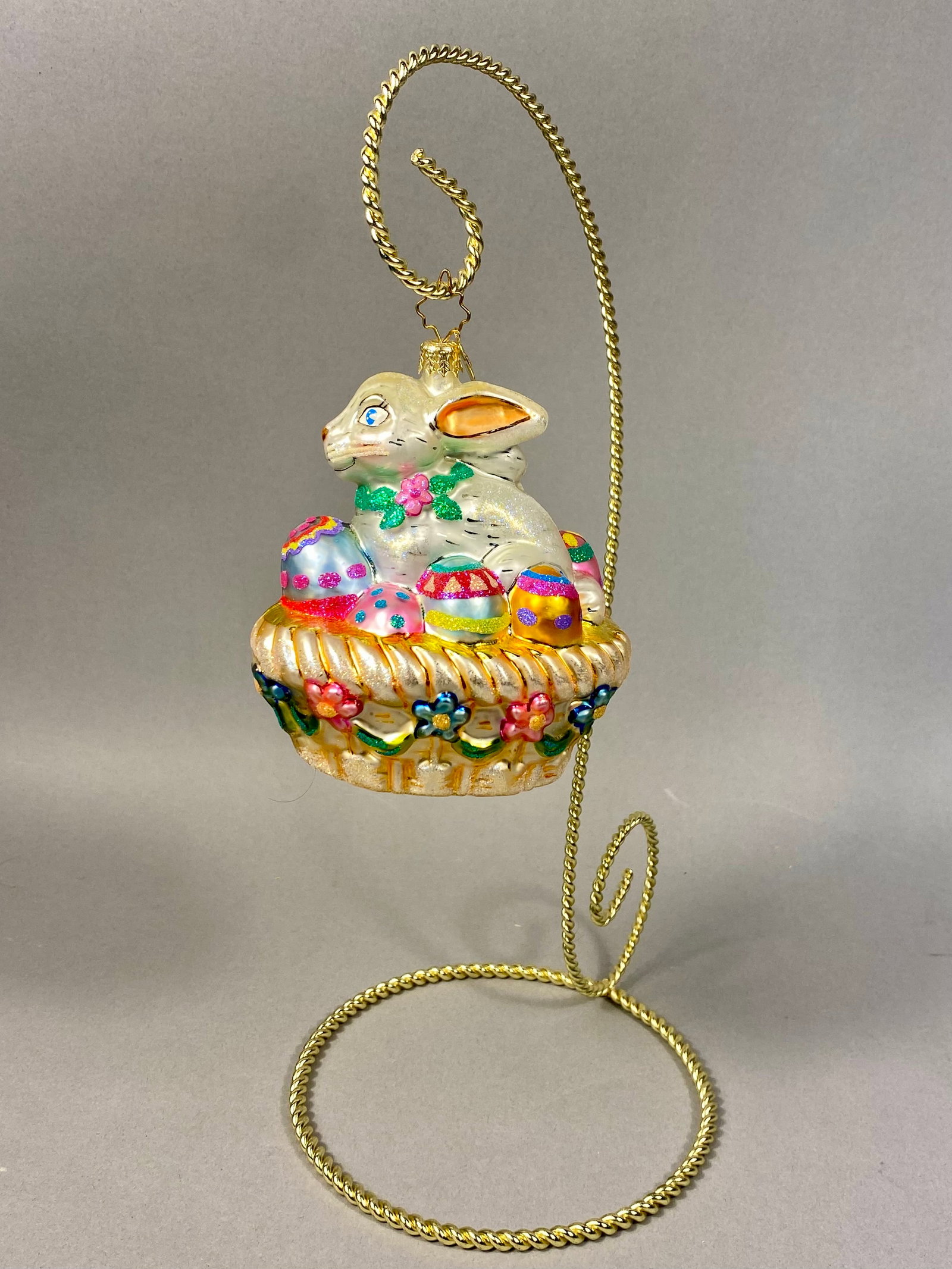 Radko Hoppy Holidays Blown Glass Easter Bunny Ornament: A mouth-blown glass figural ornament by Christopher Radko titled Hoppy Holidays. The piece depicts a pearlescent white rabbit seated within a champagne-colored basket filled with decorated Easter eggs