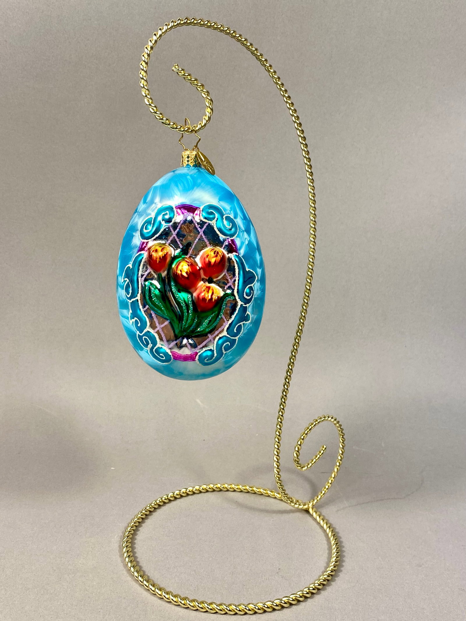 Radko Hand-Blown Glass Egg Ornament with Tulip Motif: A hand-blown glass egg ornament produced by Christopher Radko, featuring a light blue body with a crystallized feathered ice-finish effect. The piece contains a central recessed window depicting a han
