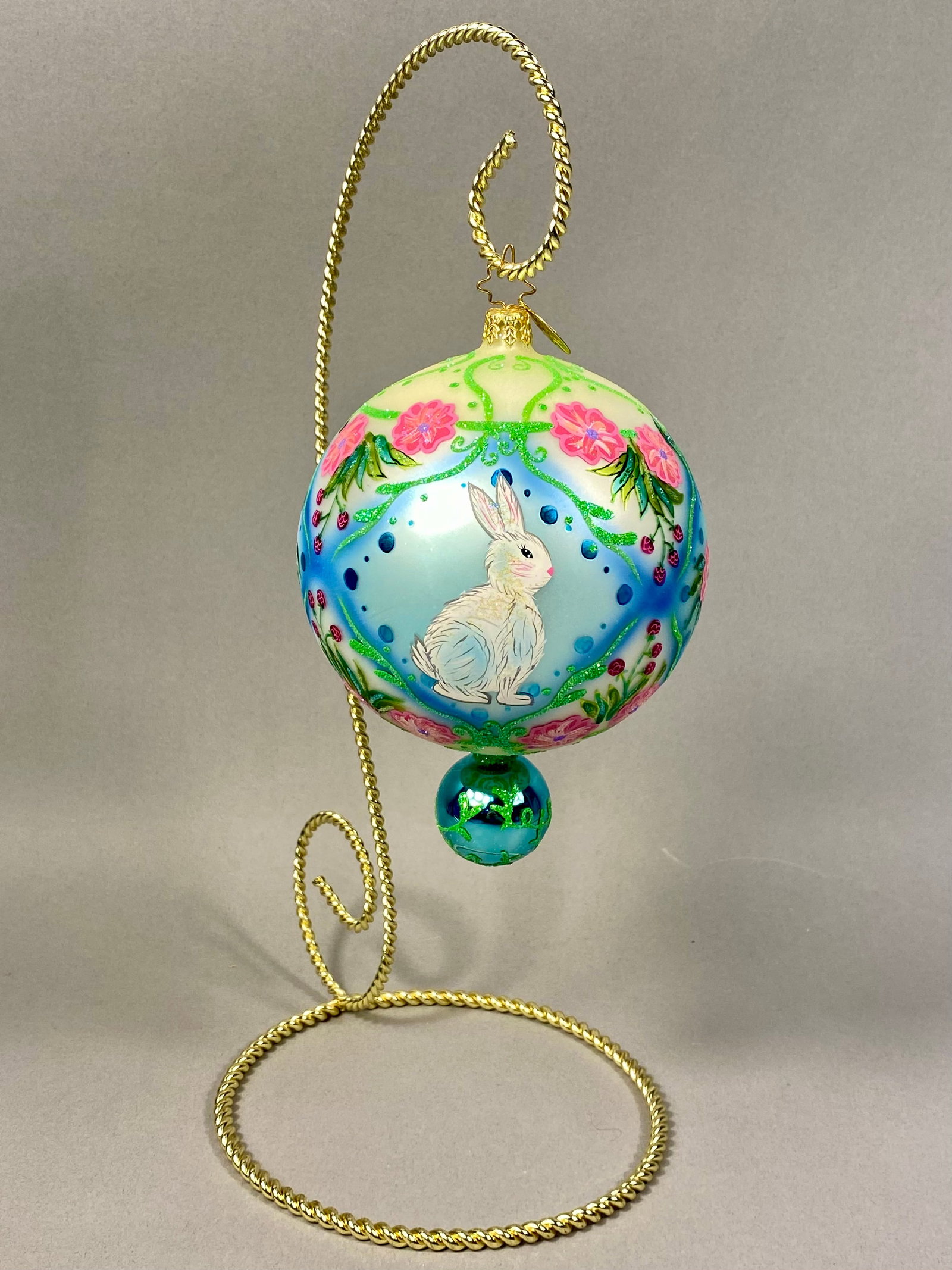 Radko Hand-Painted Glass Rabbit Ornament: This hand-blown glass ornament by Christopher Radko features a central hand-painted vignette of a white rabbit with pink inner ears and a black eye, set against a pale blue background. The ornament in