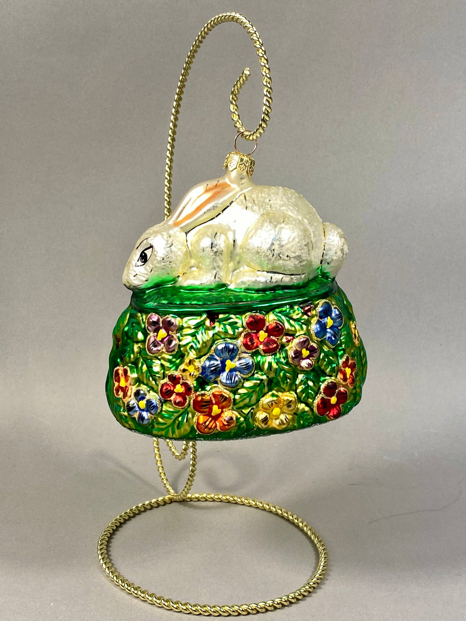Radko Healing Holiday Hare Hand-Blown Glass Ornament: This Christopher Radko hand-blown glass ornament is identified as the Healing Holiday Hare. The piece features a pearlescent white rabbit with iridescent finishes resting atop a rounded, emerald-green