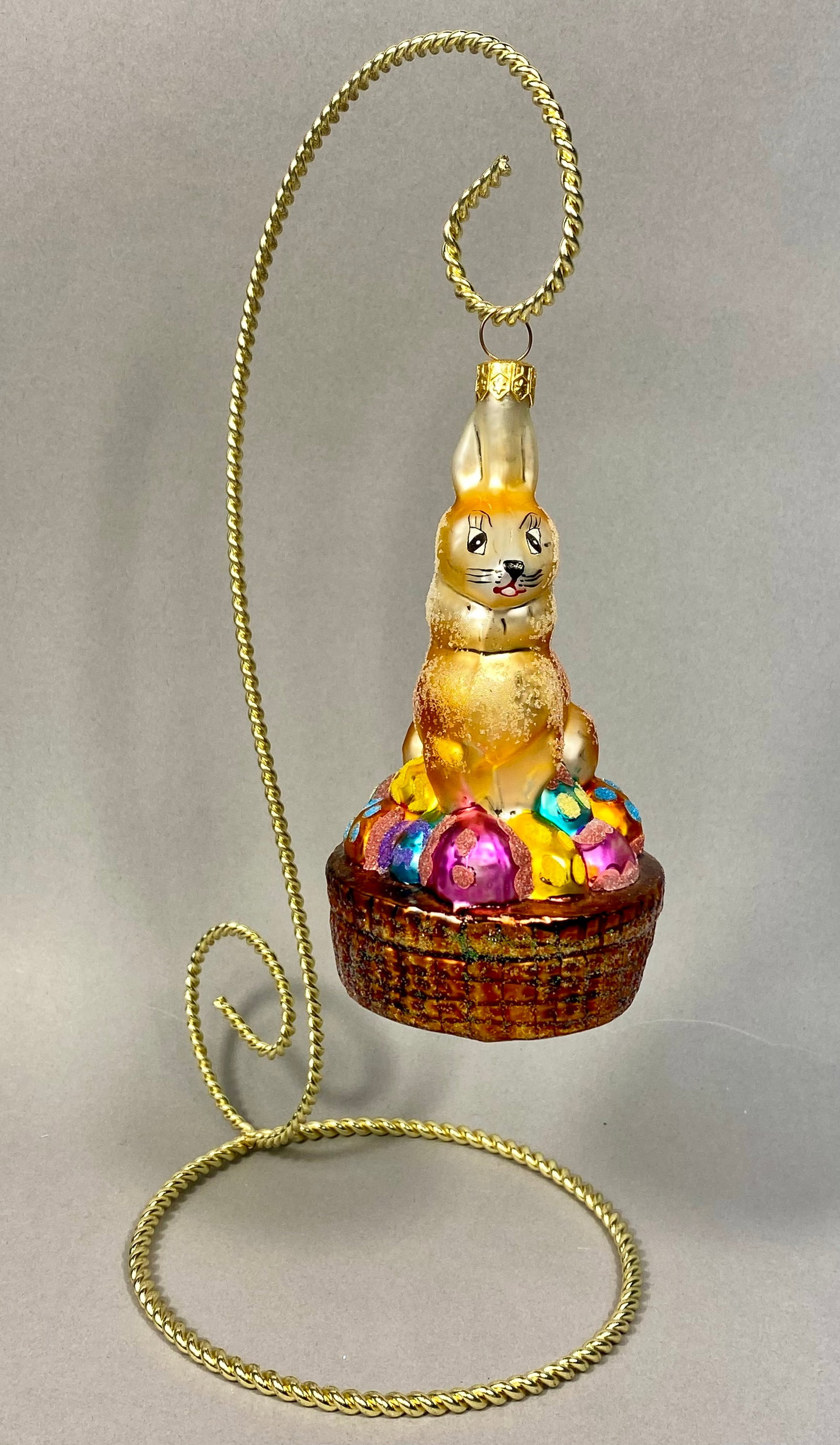 Radko Hoppy Holidays Glass Ornament: A Christopher Radko Hoppy Holidays hand-blown silvered glass Easter ornament. The figural ornament depicts a golden-amber rabbit seated atop a brown basket filled with several multi-colored Easter egg