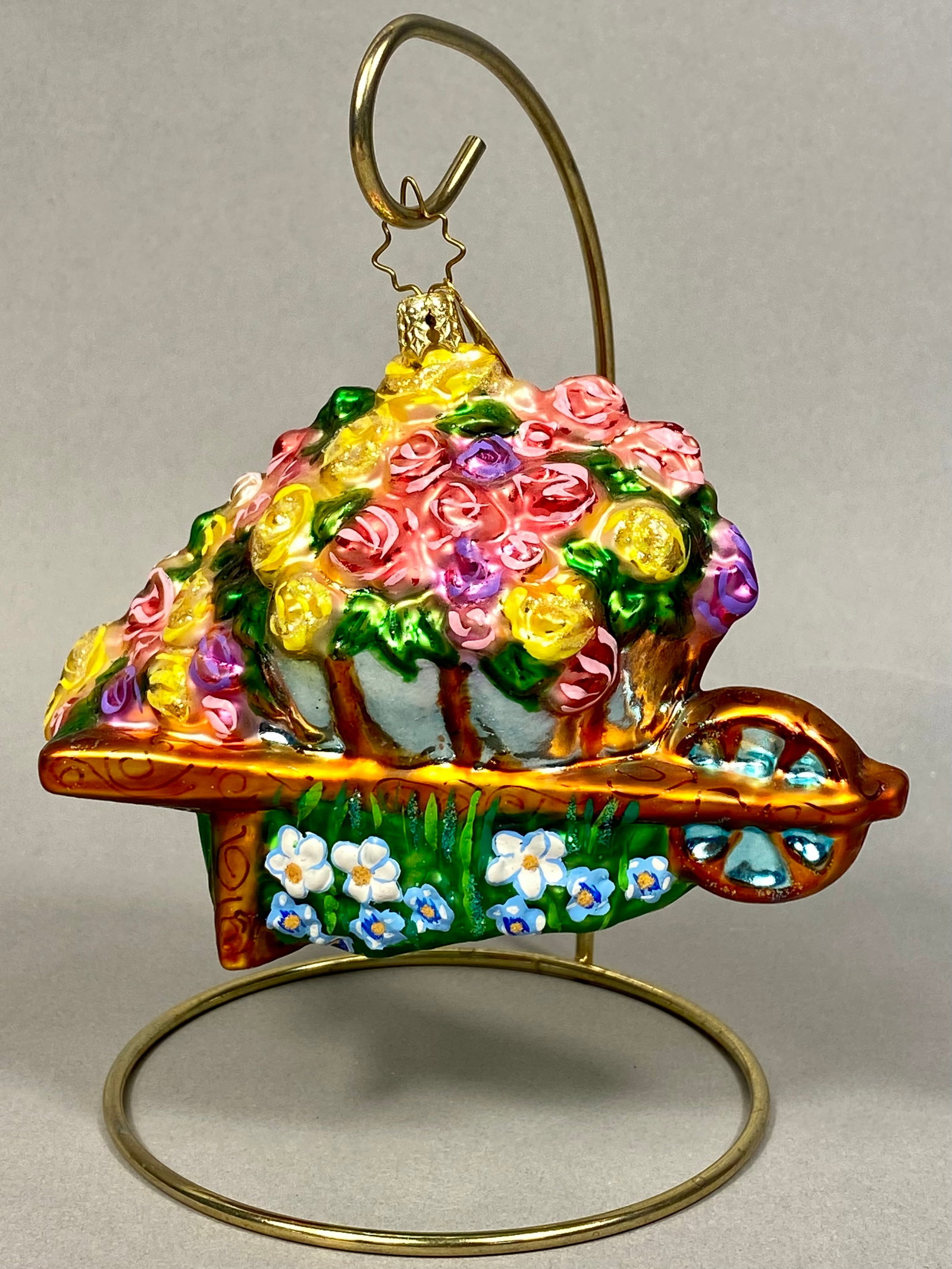 Radko Flower Wheelbarrow Mouth-Blown Glass Ornament: A Christopher Radko mouth-blown glass figural ornament depicting a wheelbarrow filled with a profusion of flowers. The ornament is fitted with an embossed gold-tone metal cap and the maker's signature