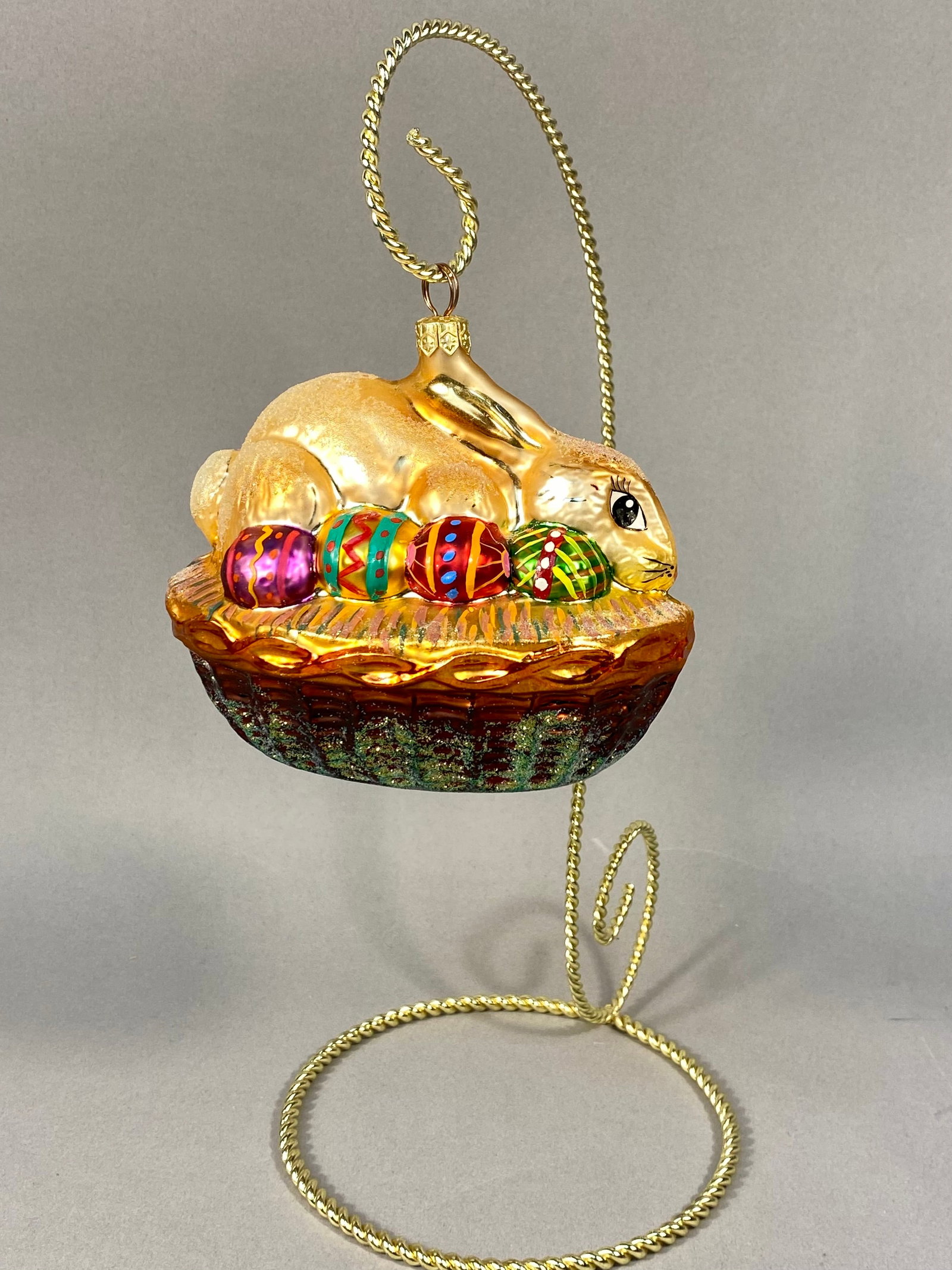 Radko Hoppy Easter Glass Ornament: This Christopher Radko glass ornament, titled Hoppy Easter, features a champagne-colored rabbit resting atop four vibrantly decorated Easter eggs within a woven-texture basket. The rabbit is finished 