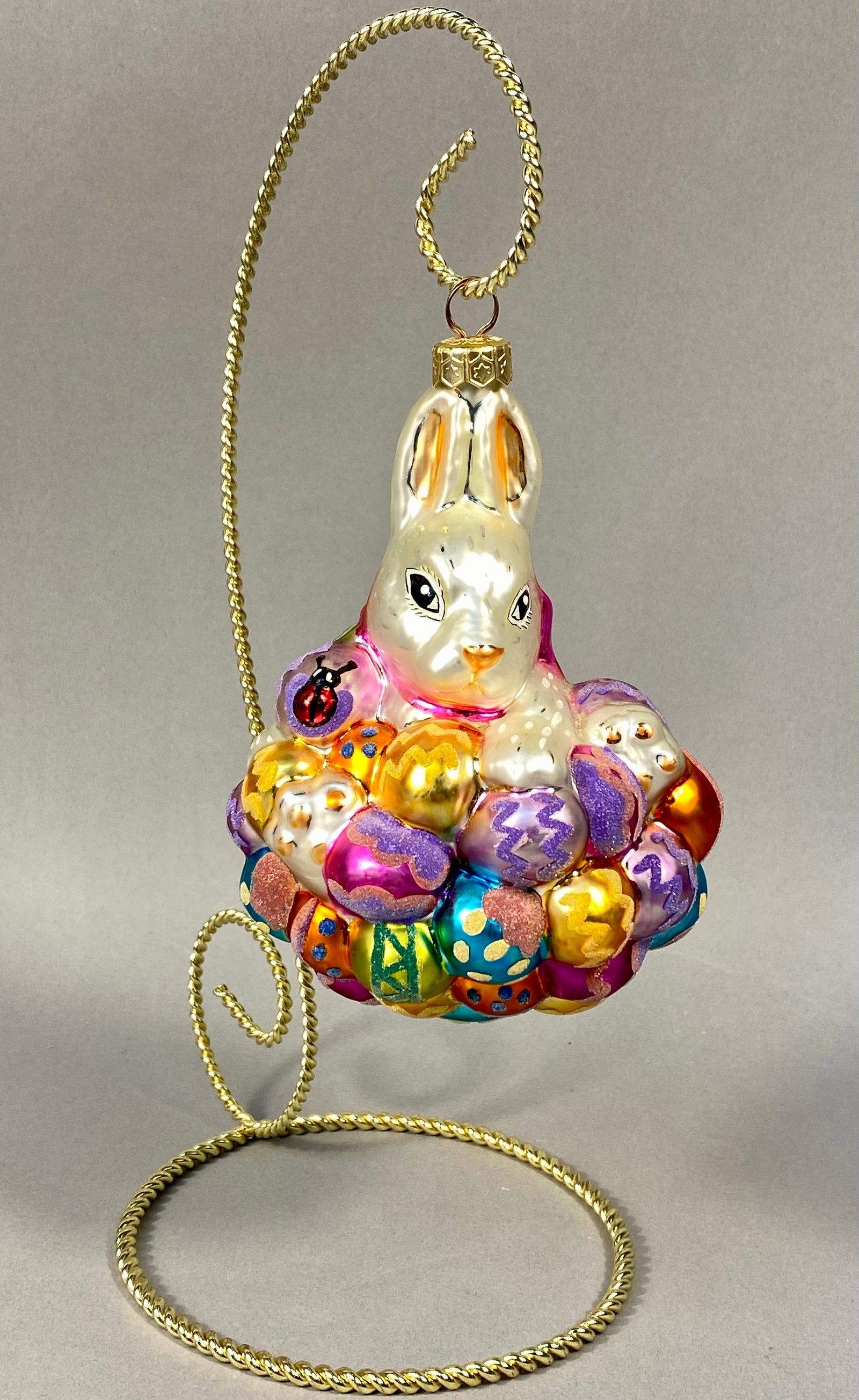 Radko Hand-Blown Glass Easter Bunny and Eggs Ornament: This hand-blown glass holiday ornament is a product of Christopher Radko. The piece features a white Easter bunny with gold-tipped ears and hand-painted facial features, nestled atop a cluster of vari