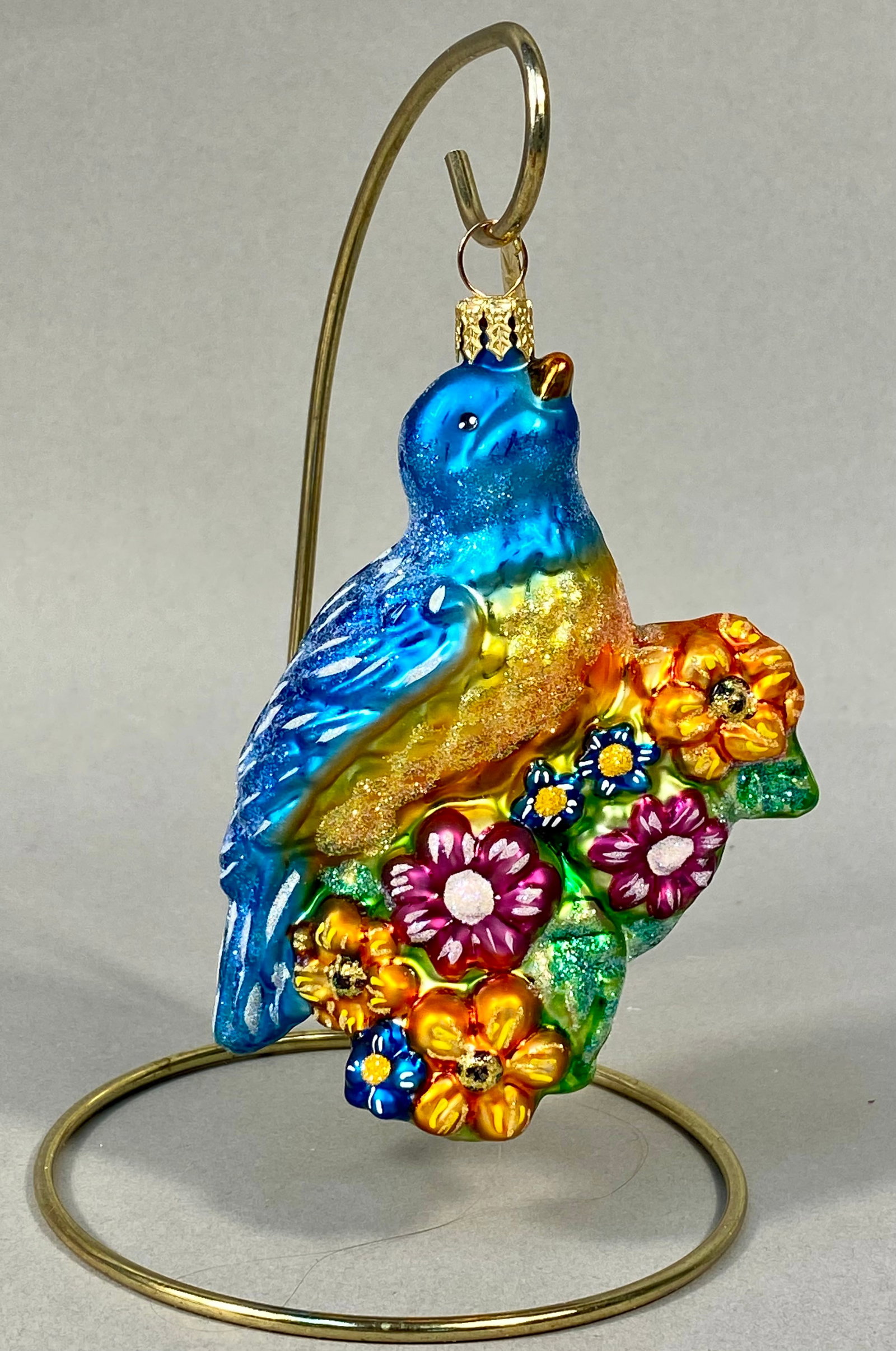 Radko Garden Guest Glass Bluebird and Flowers Ornament: A mouth-blown glass holiday ornament titled Garden Guest, produced by Christopher Radko. The ornament features a sculpted bluebird with a cerulean blue body, hand-painted wing details, and a gold-glit