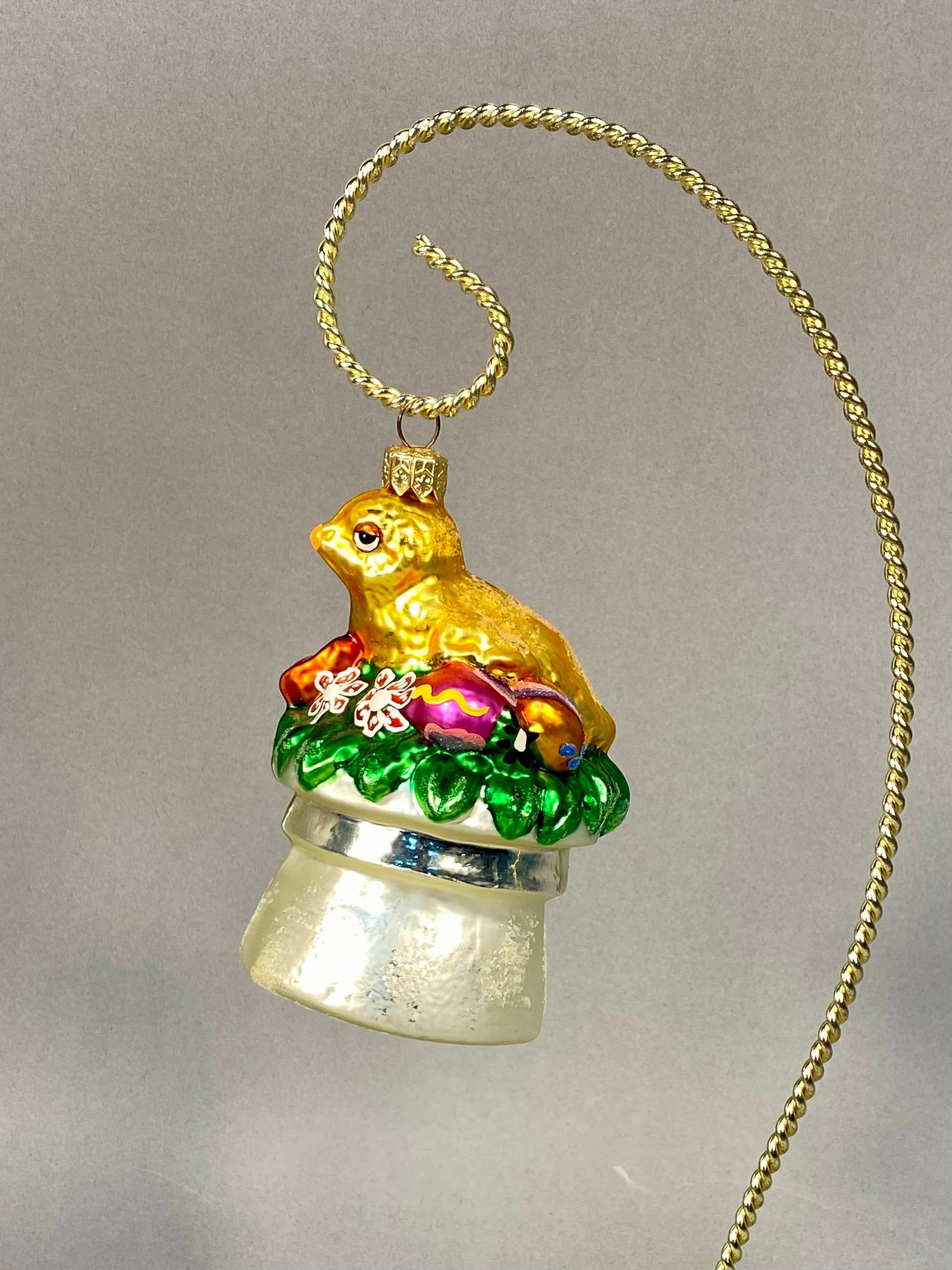 Radko Glass Easter Chick in Top Hat Ornament: A Christopher Radko mouth-blown glass ornament. The design features a metallic gold-toned chick figure in a seated position resting on a bed of green foliage. The ornament is hand-painted with lacquer