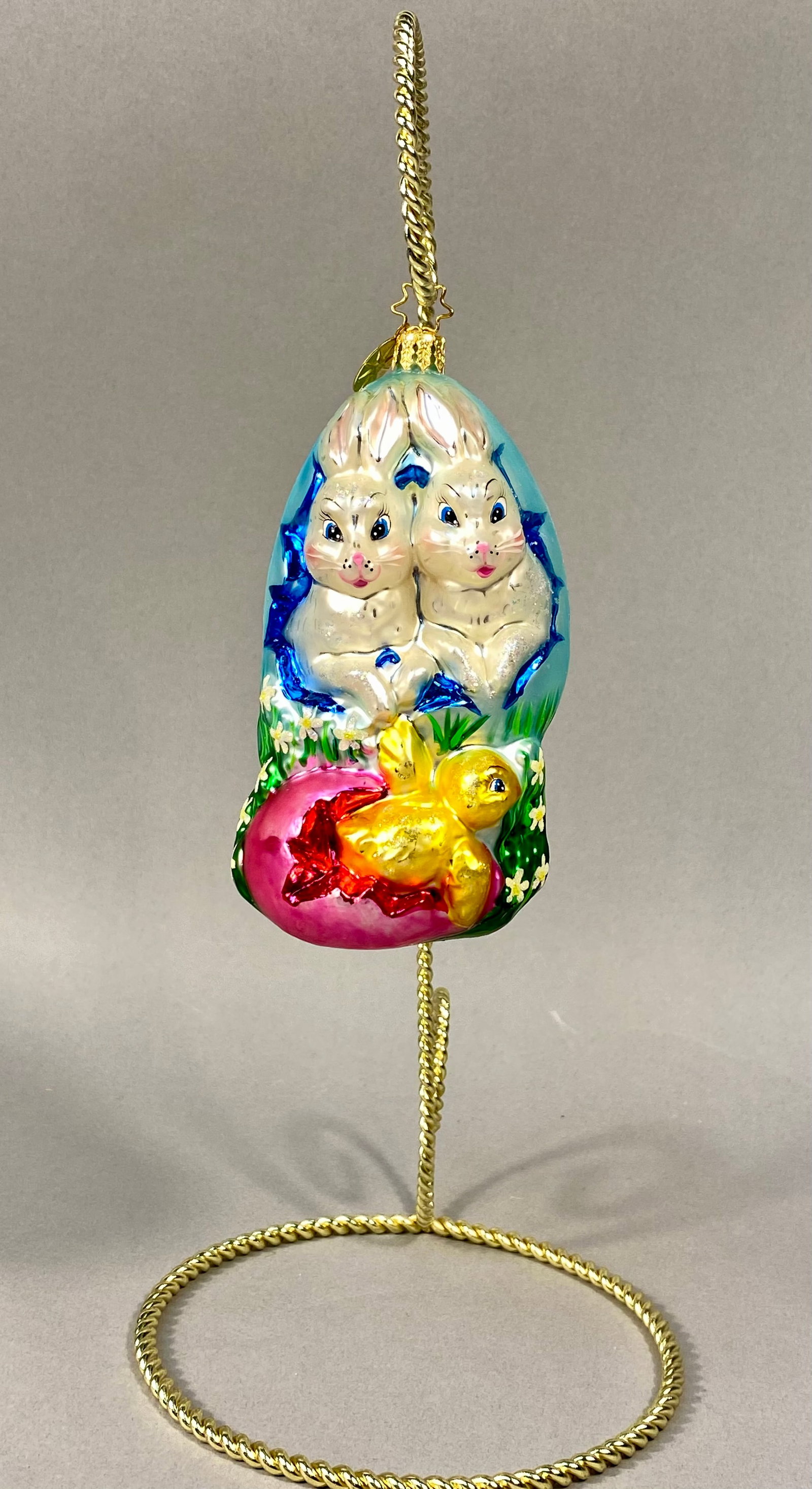 Radko Hoppy Holidays Glass Easter Ornament: A mouth-blown glass holiday ornament by designer Christopher Radko, specifically the Hoppy Holidays pattern. The ornament depicts two white rabbits with blue eyes emerging from a large, light blue cra