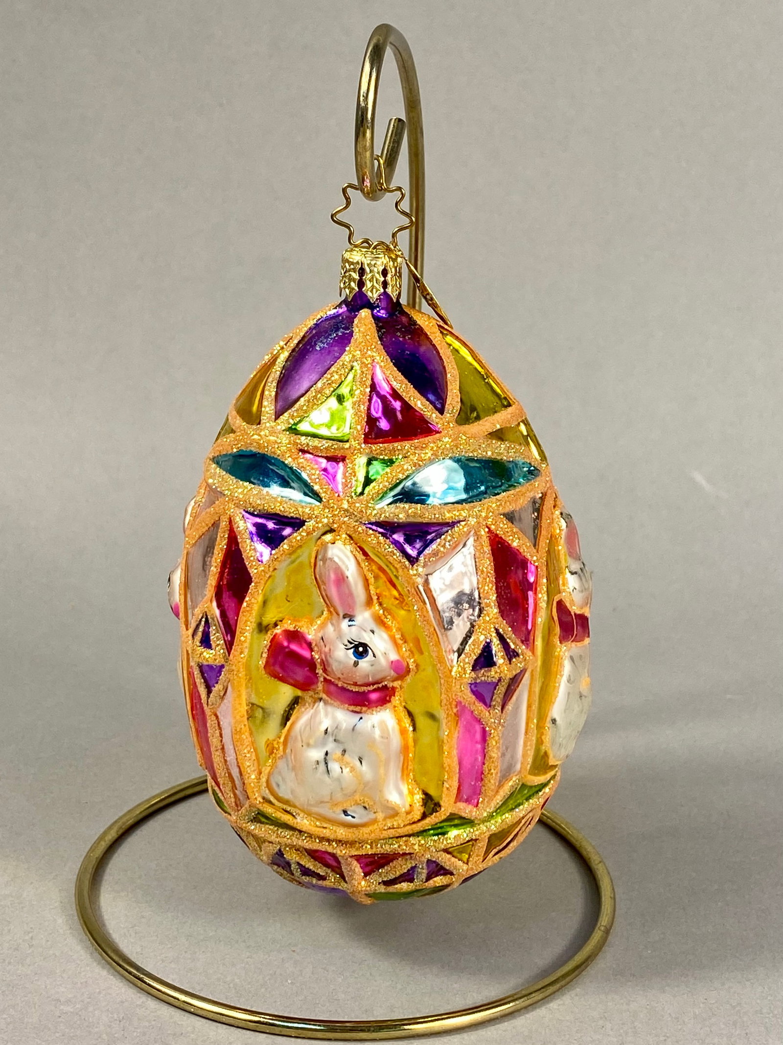 Radko Stained Glass Style Rabbit Motif Glass Egg Ornament: This item is a mouth-blown and hand-painted glass Easter ornament produced by Christopher Radko. The egg-shaped ornament features a stained glass aesthetic with multiple colorful panels in translucent