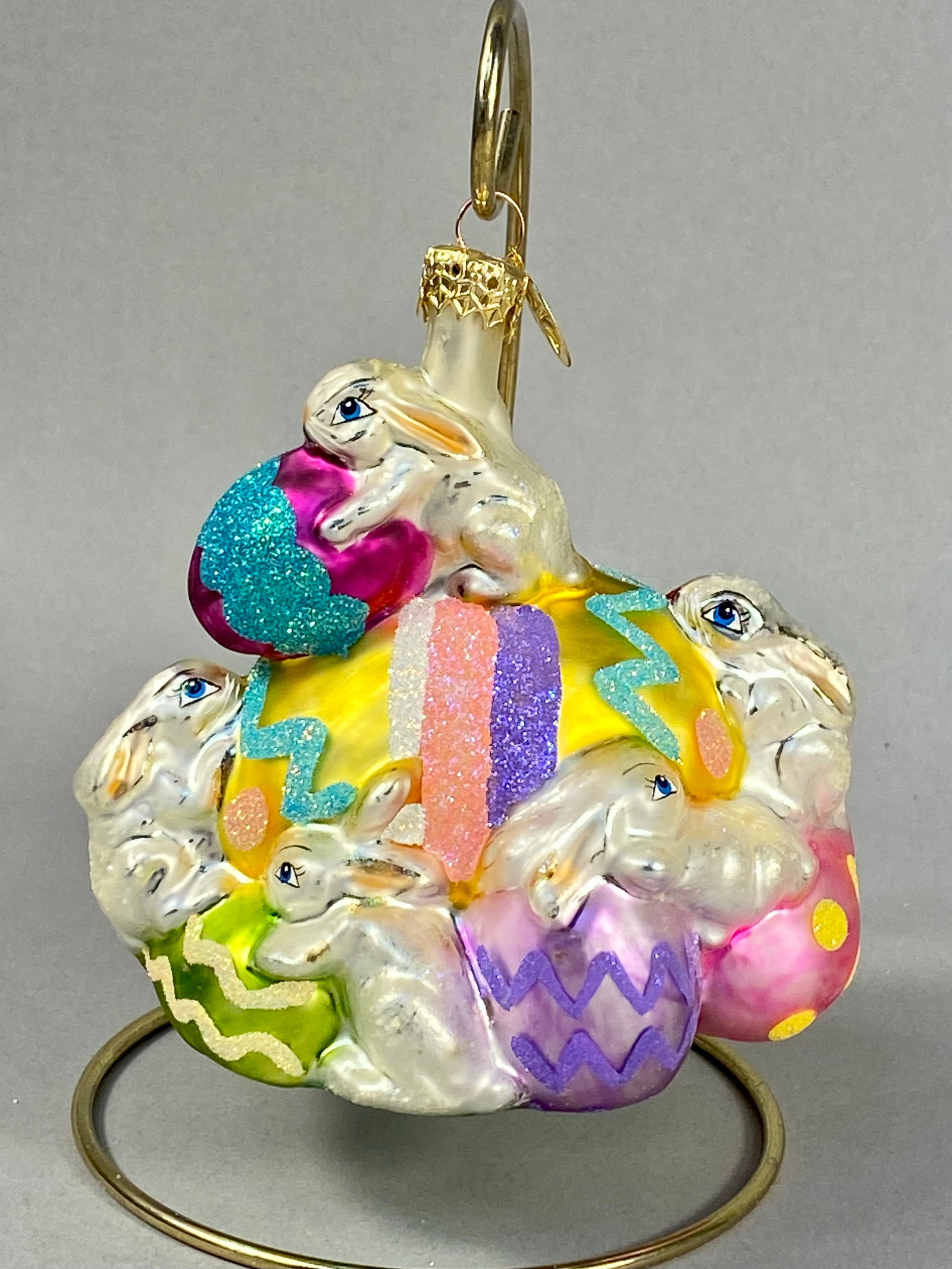Radko Hoppy Easter Glass Holiday Ornament: Christopher Radko Hoppy Easter mouth-blown glass holiday ornament. The design features a large central yellow egg decorated with vertical glittered stripes in purple, pink, and white, as well as a tea