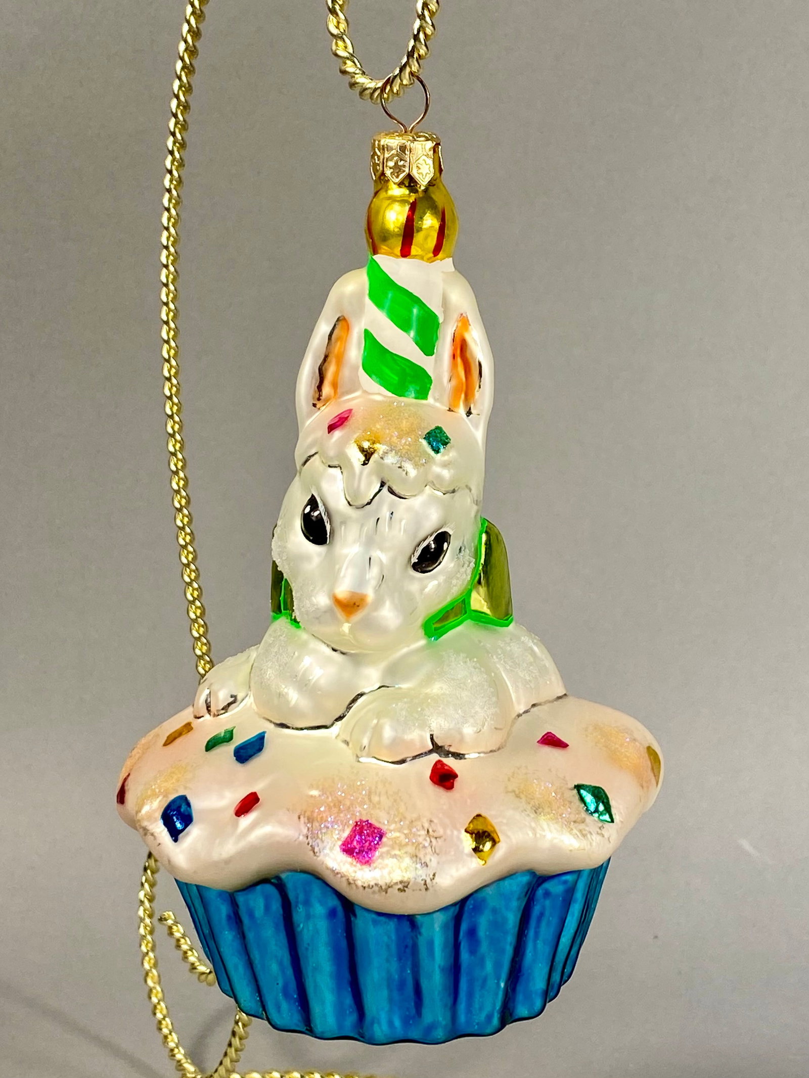 Radko Glass Rabbit on Cupcake Holiday Ornament: A hand-blown glass ornament manufactured by Christopher Radko, featuring a whimsical design of a white rabbit sitting atop a frosted cupcake. The reverse side of the cupcake frosting features a small,