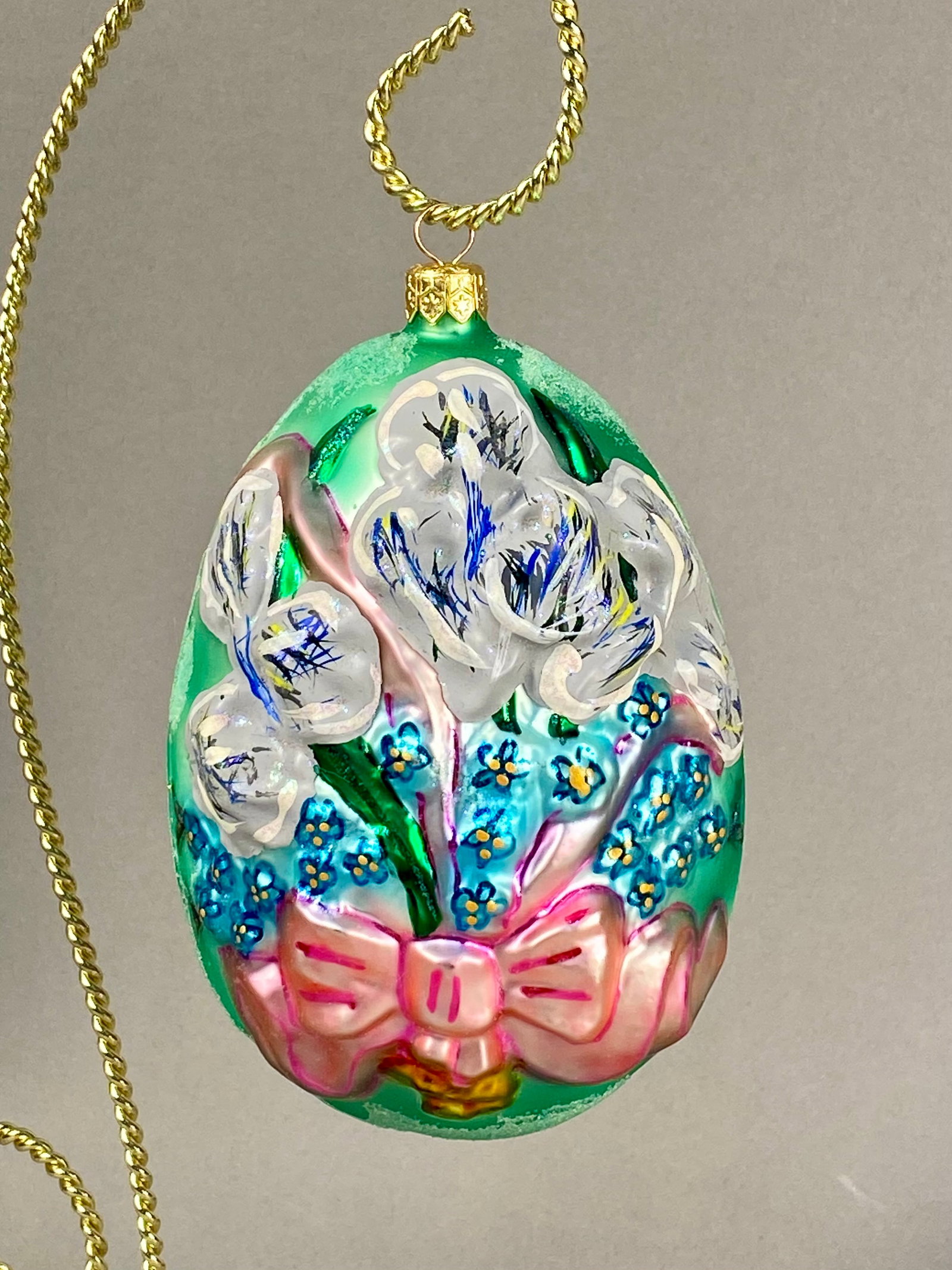 Radko Hand-Painted Glass Floral Egg Ornament: The piece features a vibrant mint green ground decorated with a hand-painted floral bouquet. The base of the bouquet is finished with a large, molded pink bow accented with darker pink lines. The orna