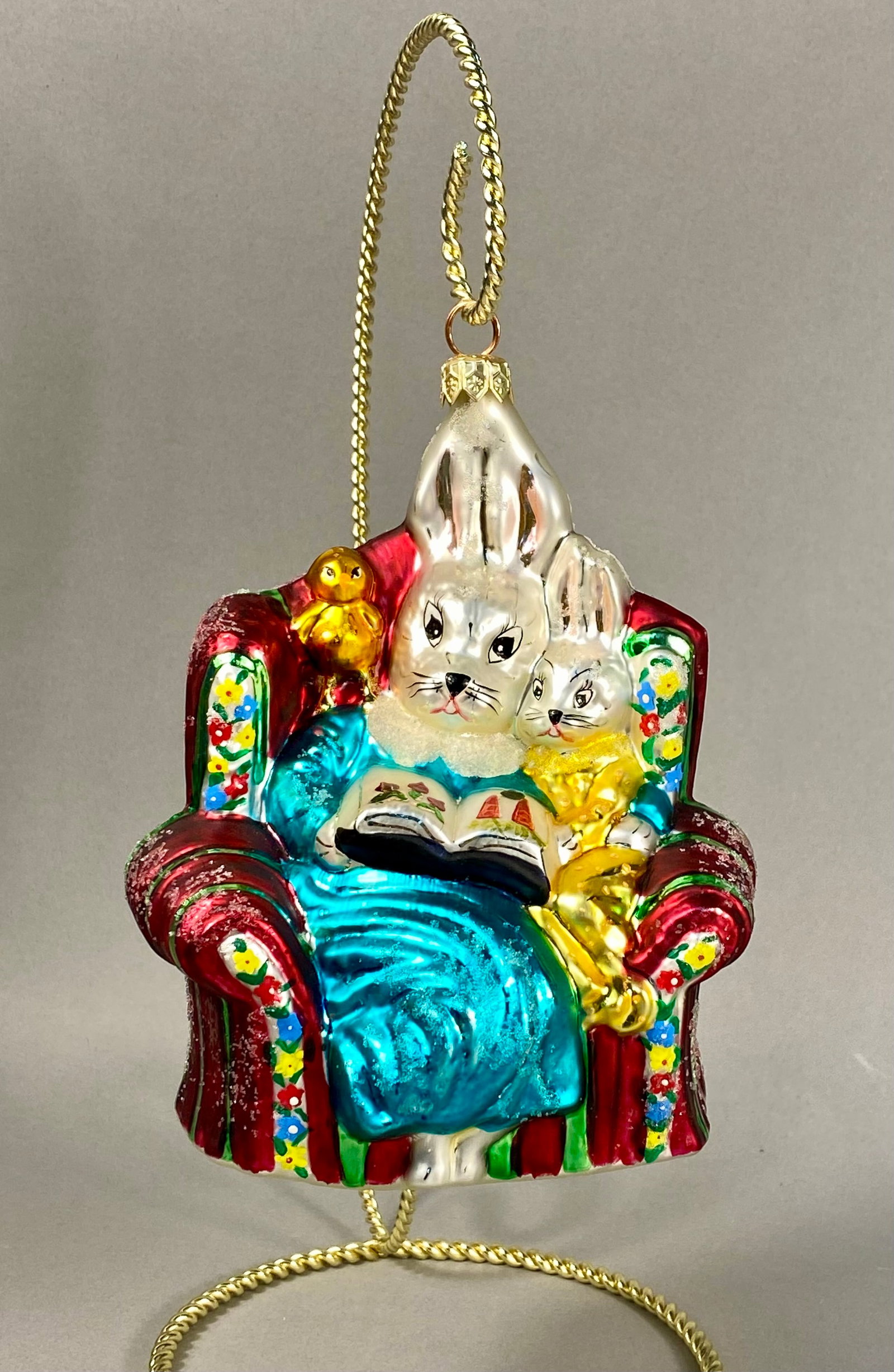 Radko 1998 Bedtime Story Glass Ornament: This hand-blown glass ornament by Christopher Radko, titled Bedtime Story and released in 1998, depicts a whimsical scene of two white rabbits. A small yellow chick is perched on the upper shoulder of