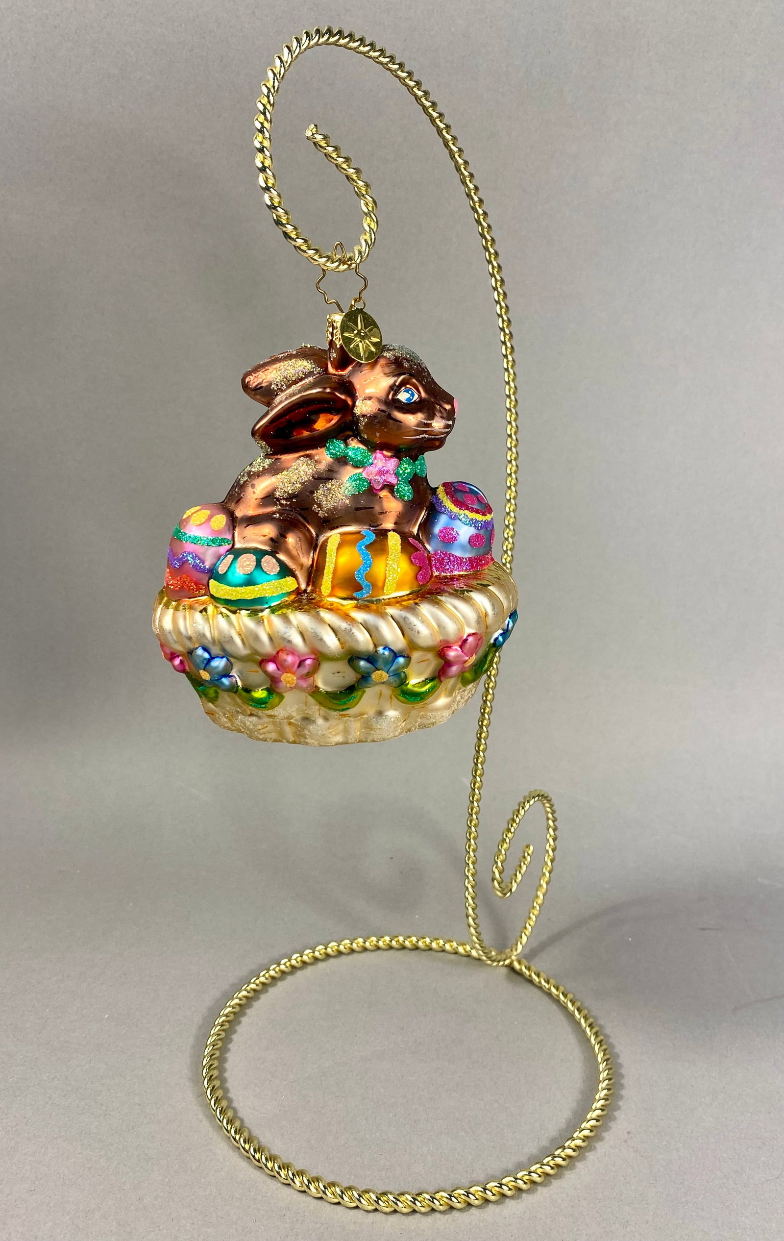 Radko Hoppy Easter Glass Ornament: This hand-blown glass holiday ornament is titled Hoppy Easter, produced by Christopher Radko. The piece depicts a seated brown rabbit with blue eyes and a pink nose, wearing a green glittered collar a